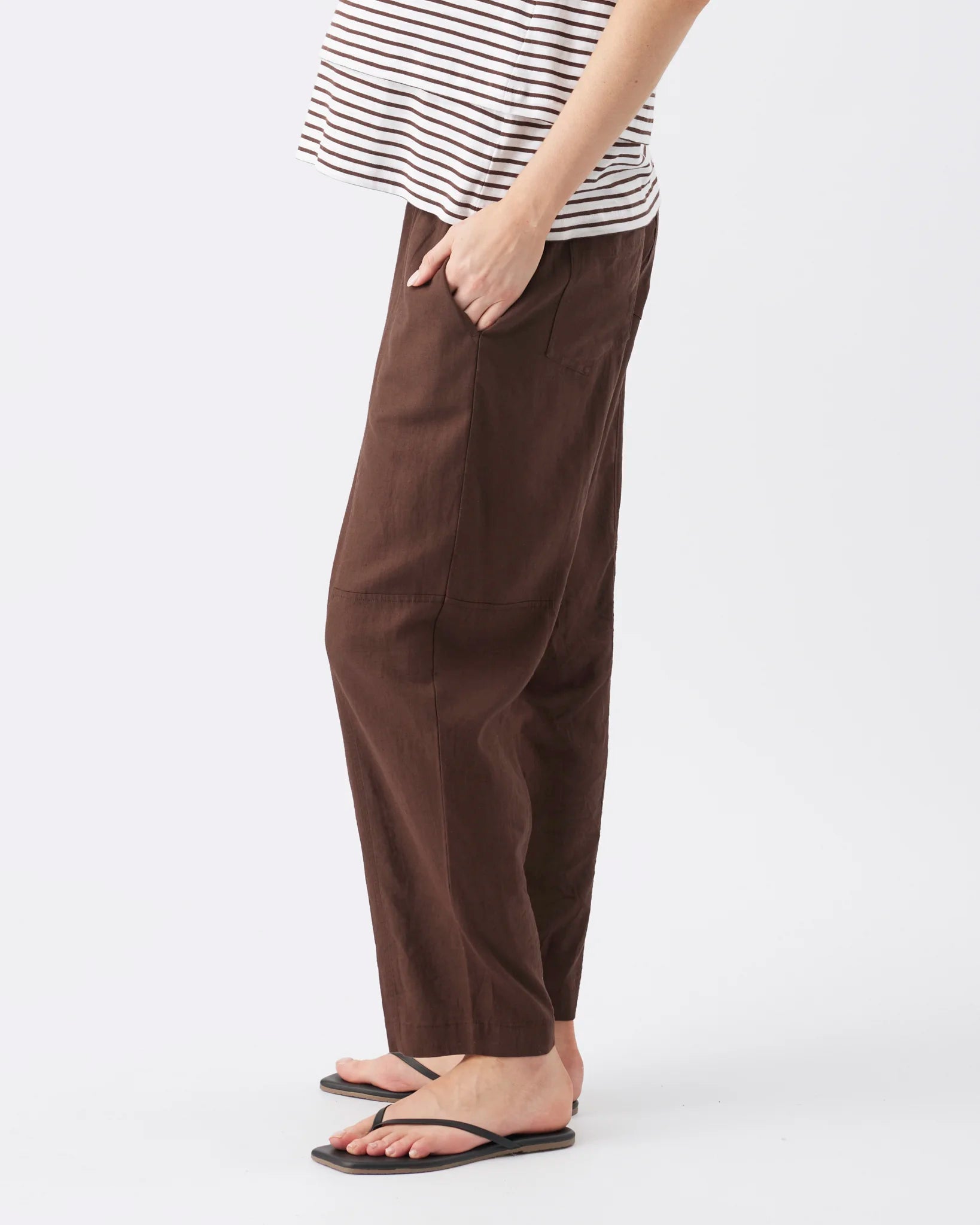 A person wearing loose brown wide-leg pants with a striped shirt and black flip-flops, shown from the waist down against a light gray studio background.
