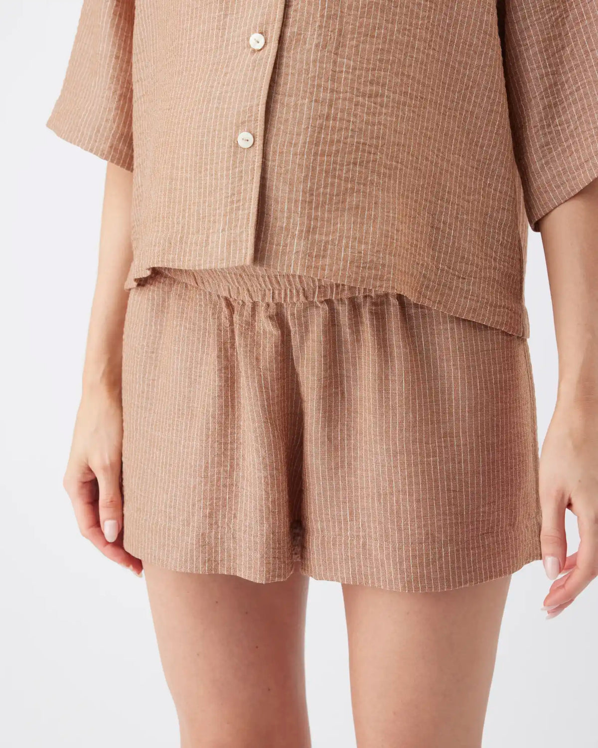 A close-up of a person wearing a matching two-piece lounge set in a textured beige fabric, featuring a short-sleeve button-front top and high-waisted shorts.
