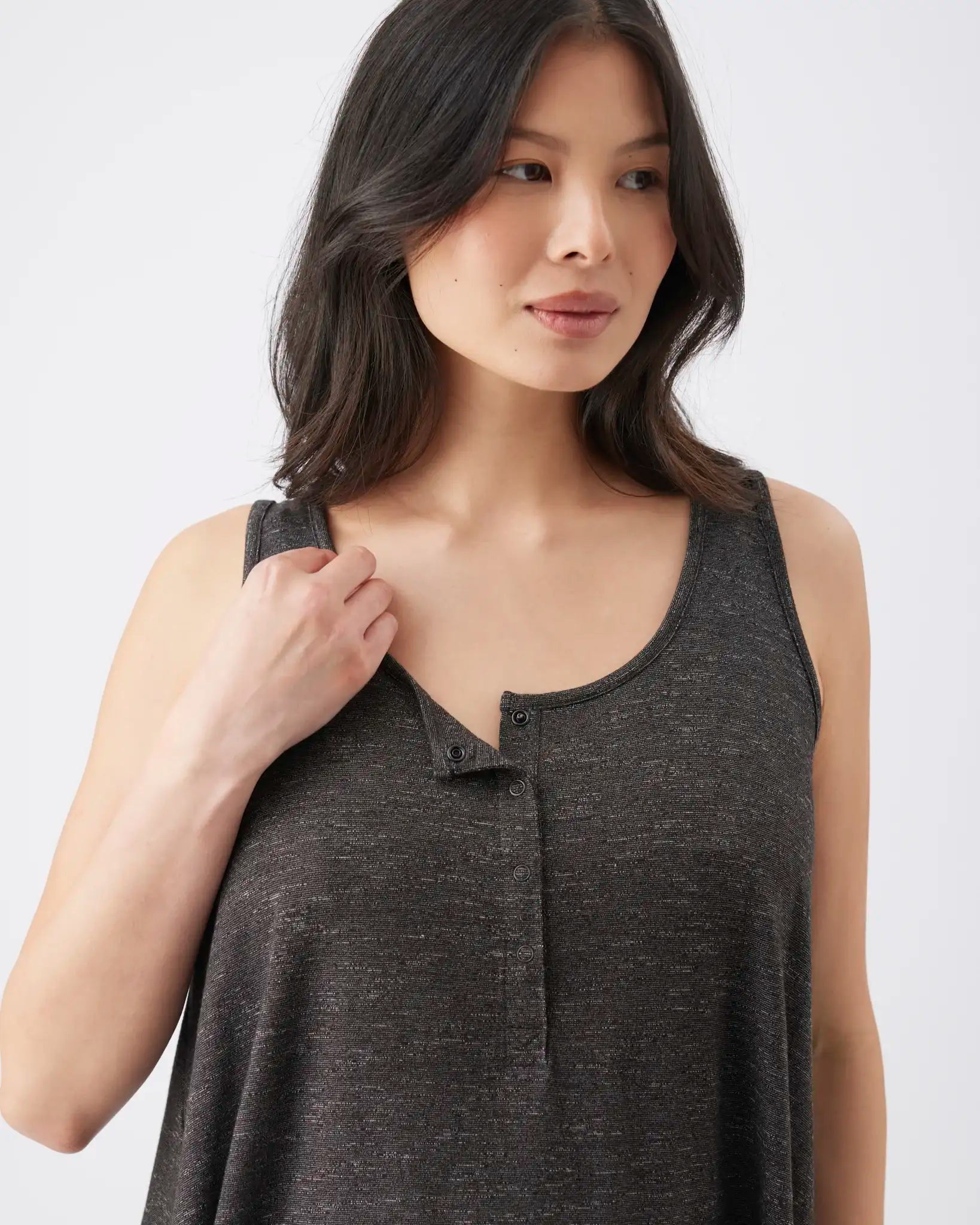 A woman wearing a dark gray heathered sleeveless henley tank top, holding the shoulder strap with one hand against a plain light gray background.