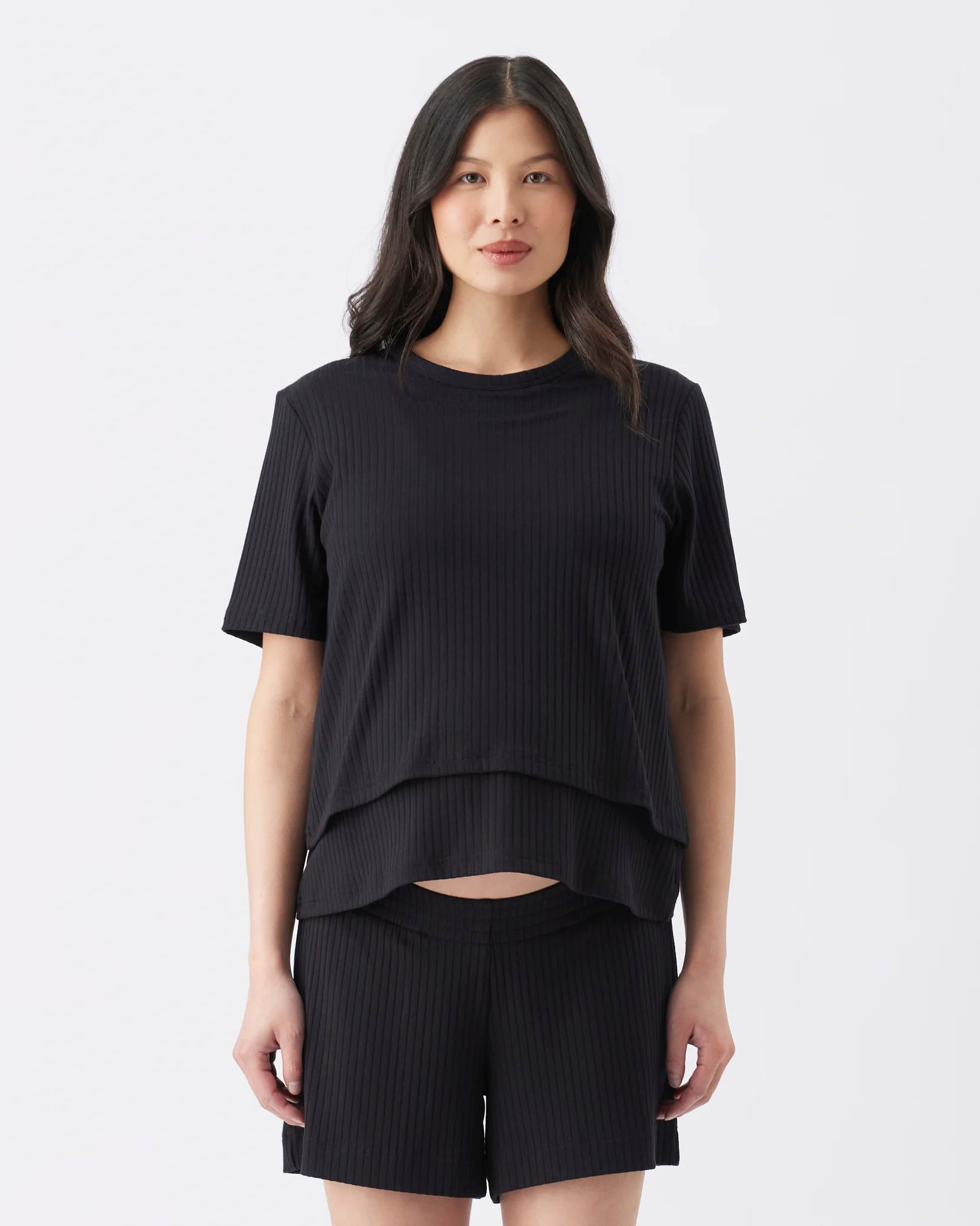 A woman modeling a black ribbed short-sleeve crop top and matching black ribbed shorts against a plain light gray studio background.