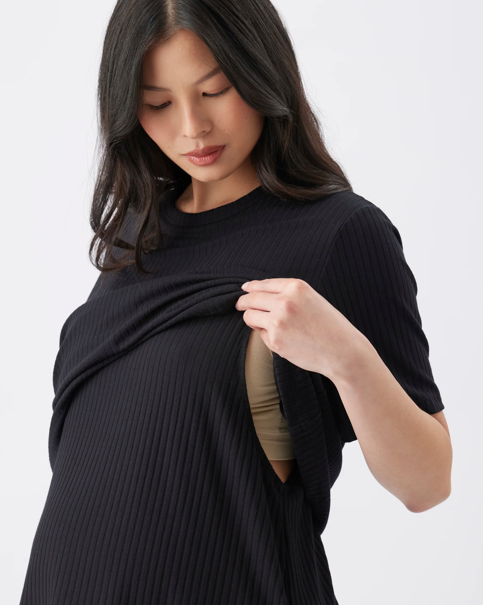 A pregnant woman wearing a black ribbed short-sleeve maternity top, lifting the fabric to reveal a beige/nude undergarment underneath, set against a plain light gray background.