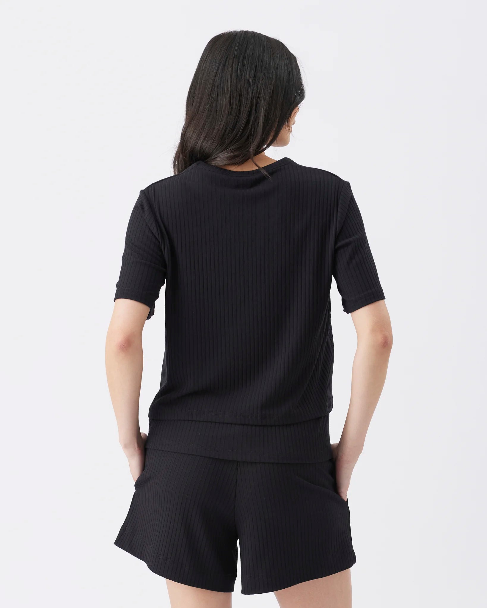 A woman shown from the back wearing a black short-sleeve ribbed knit top and matching black shorts against a plain light gray studio background.