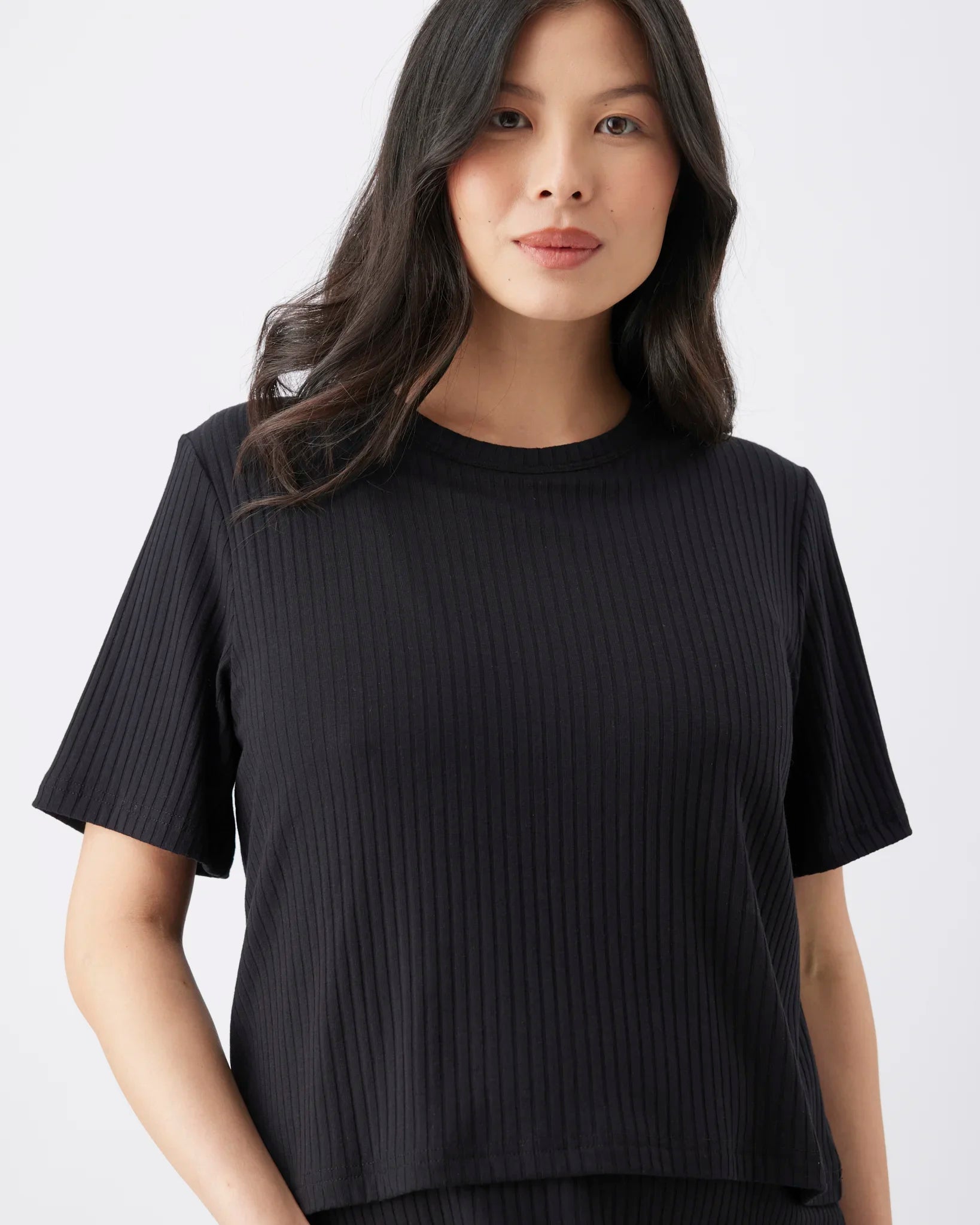 A woman wearing a black short-sleeve ribbed knit top against a plain light gray studio background.