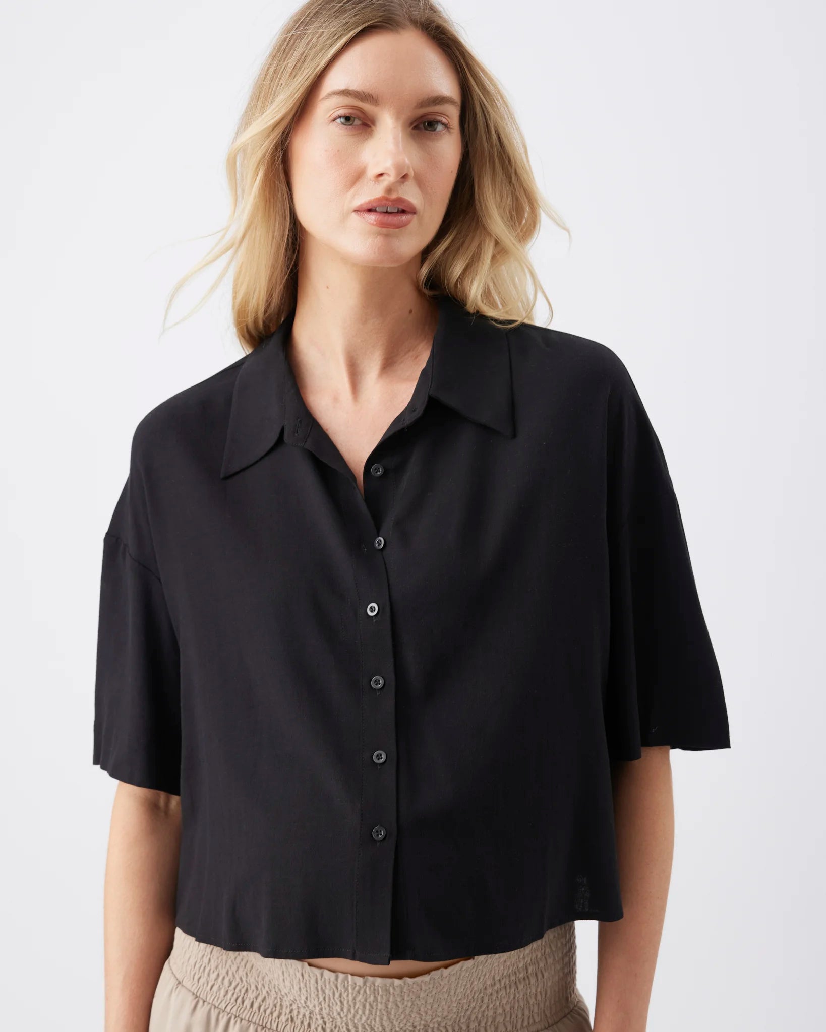 A woman wearing a black short-sleeve button-up shirt with a pointed collar, shown from the waist up against a plain light gray studio background.