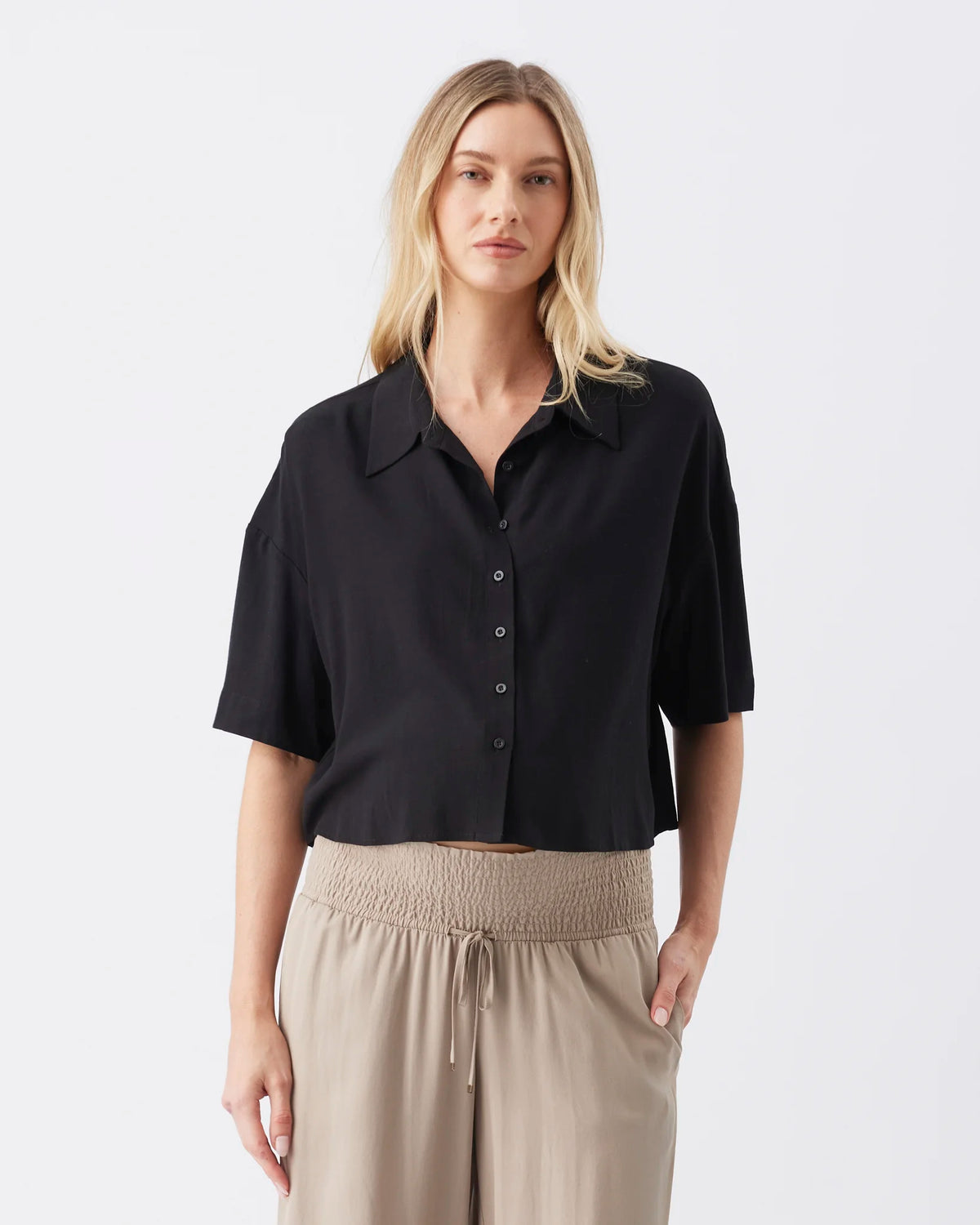 A woman modeling a black short-sleeve button-up shirt with a relaxed collar and beige drawstring trousers, shot against a clean light gray studio background.