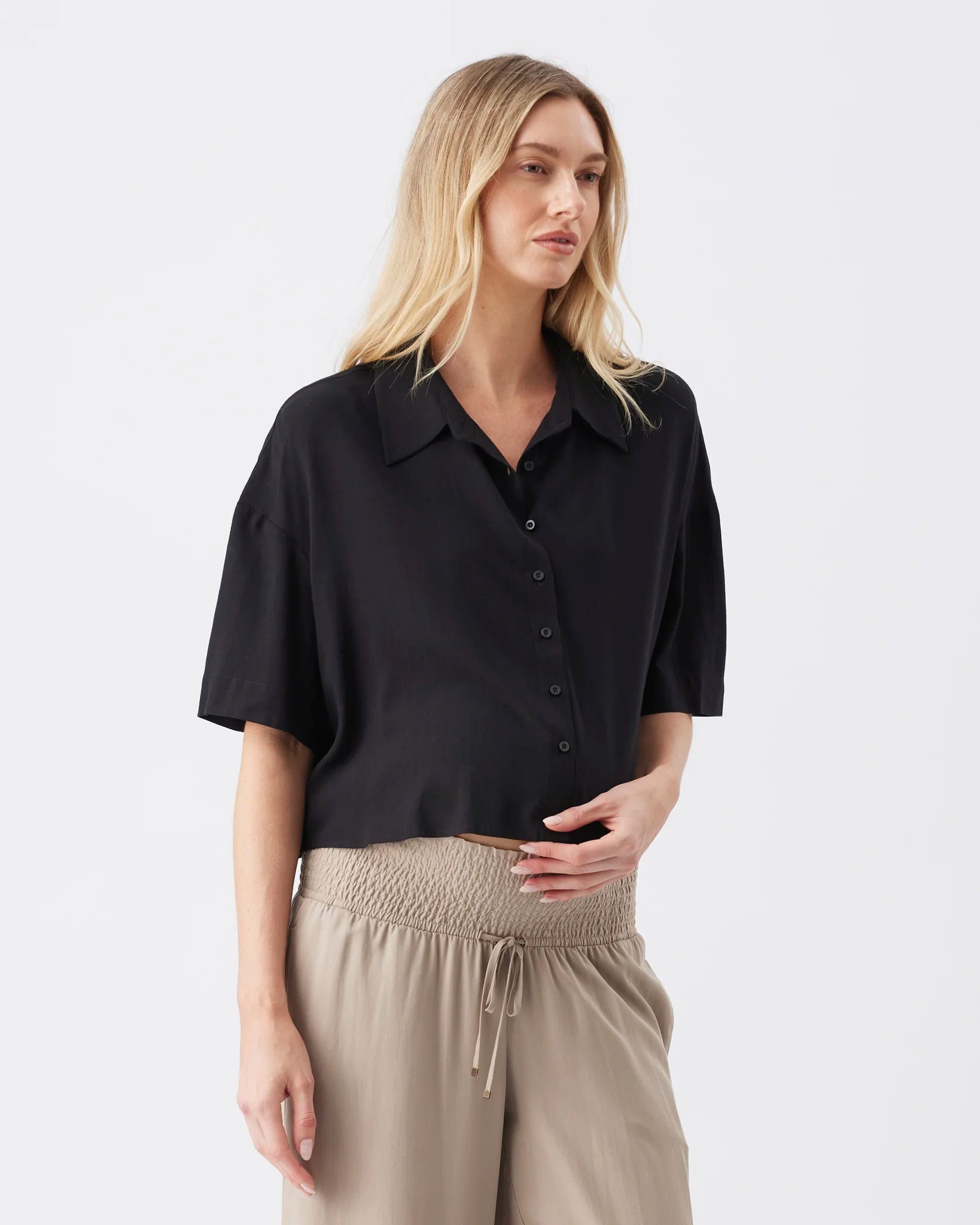 A woman modeling a black short-sleeve button-up shirt with a relaxed, oversized fit, paired with beige drawstring trousers, against a plain light gray studio background.