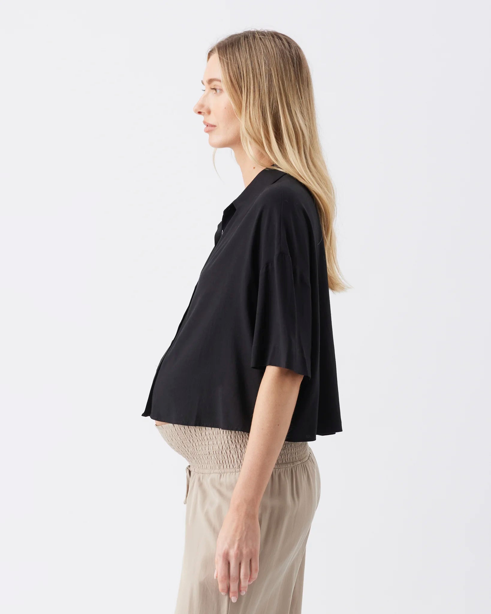 A woman shown in side profile wearing a black short-sleeve cropped button-up shirt and light beige pants against a plain light gray studio background.