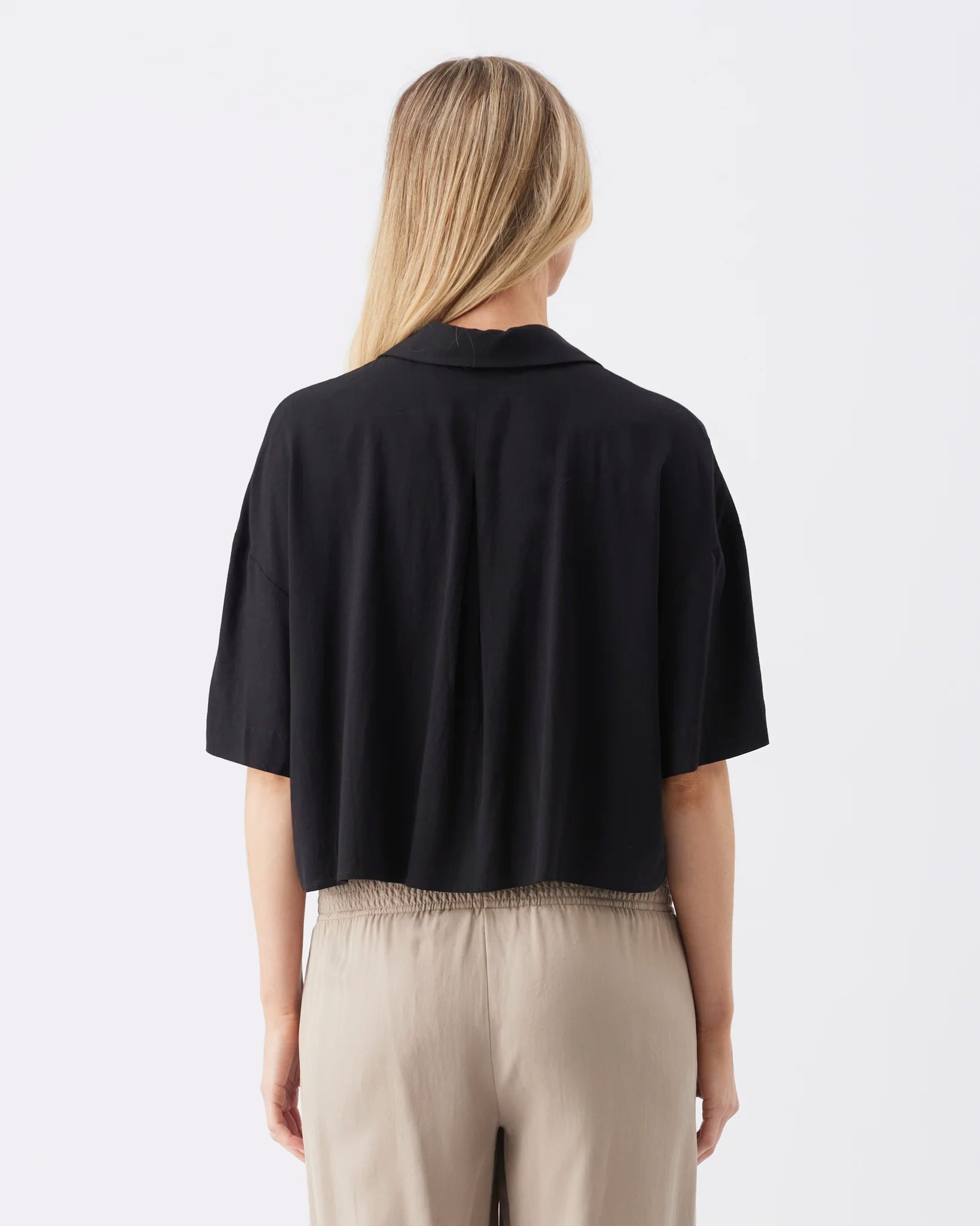A woman viewed from the back wearing a black short-sleeve collared top and light beige trousers against a plain white studio background.