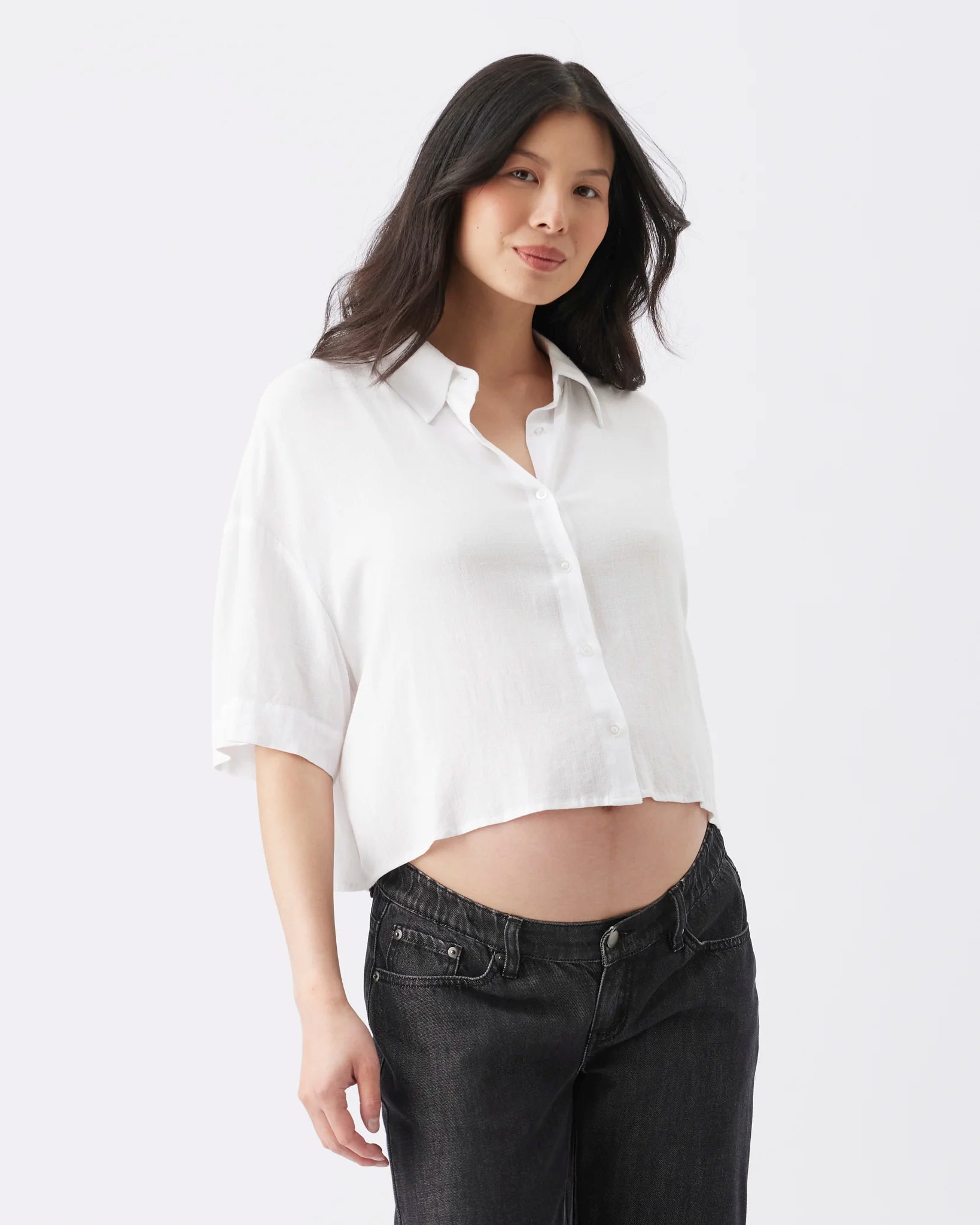 A woman modeling a white cropped button-up shirt with three-quarter sleeves and dark gray jeans against a plain white studio background.