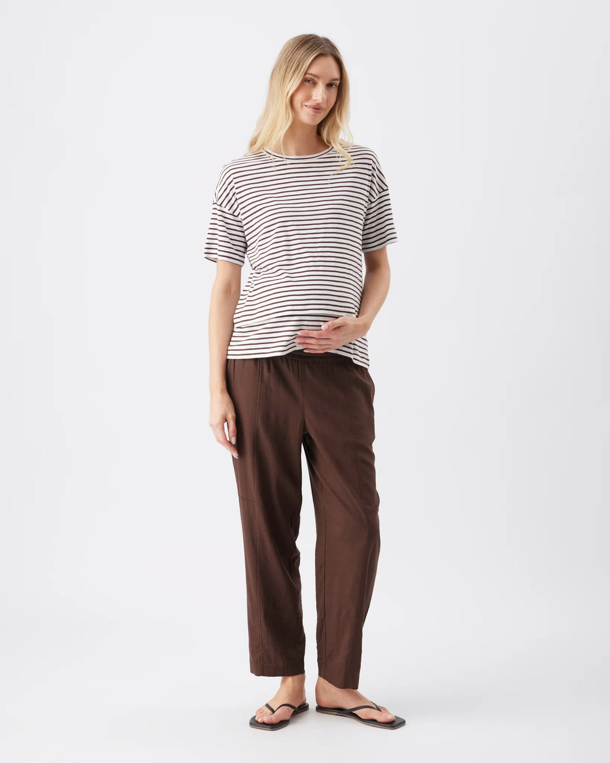 A woman modeling a short-sleeve striped t-shirt and dark brown wide-leg pants against a plain light gray studio background.
