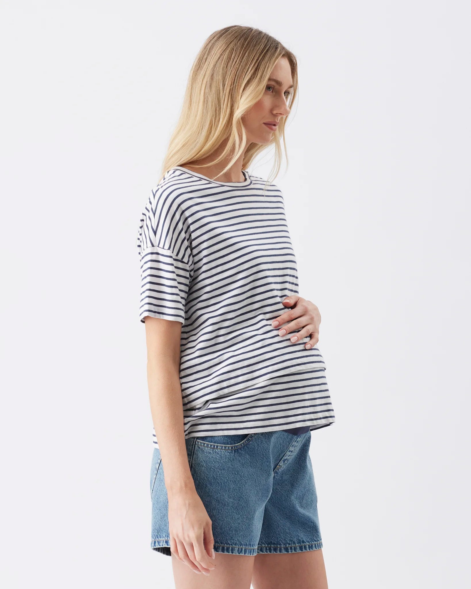 A woman wearing a black and white horizontally striped short-sleeve t-shirt and blue denim shorts, shown from a side profile against a plain white studio background.