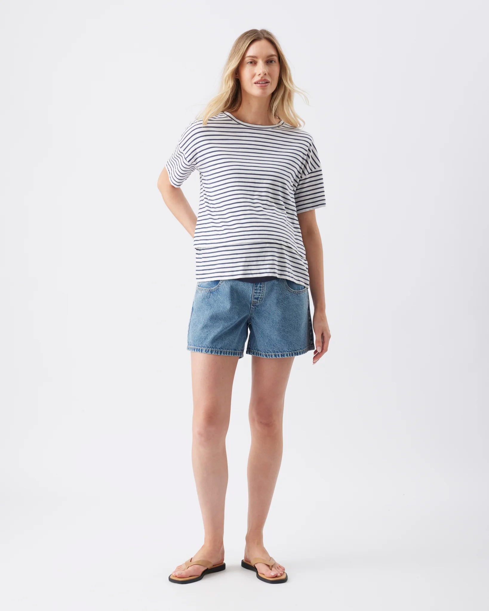 A woman modeling a short-sleeve striped t-shirt in navy and white, paired with blue denim shorts and tan sandals, against a plain white studio background.