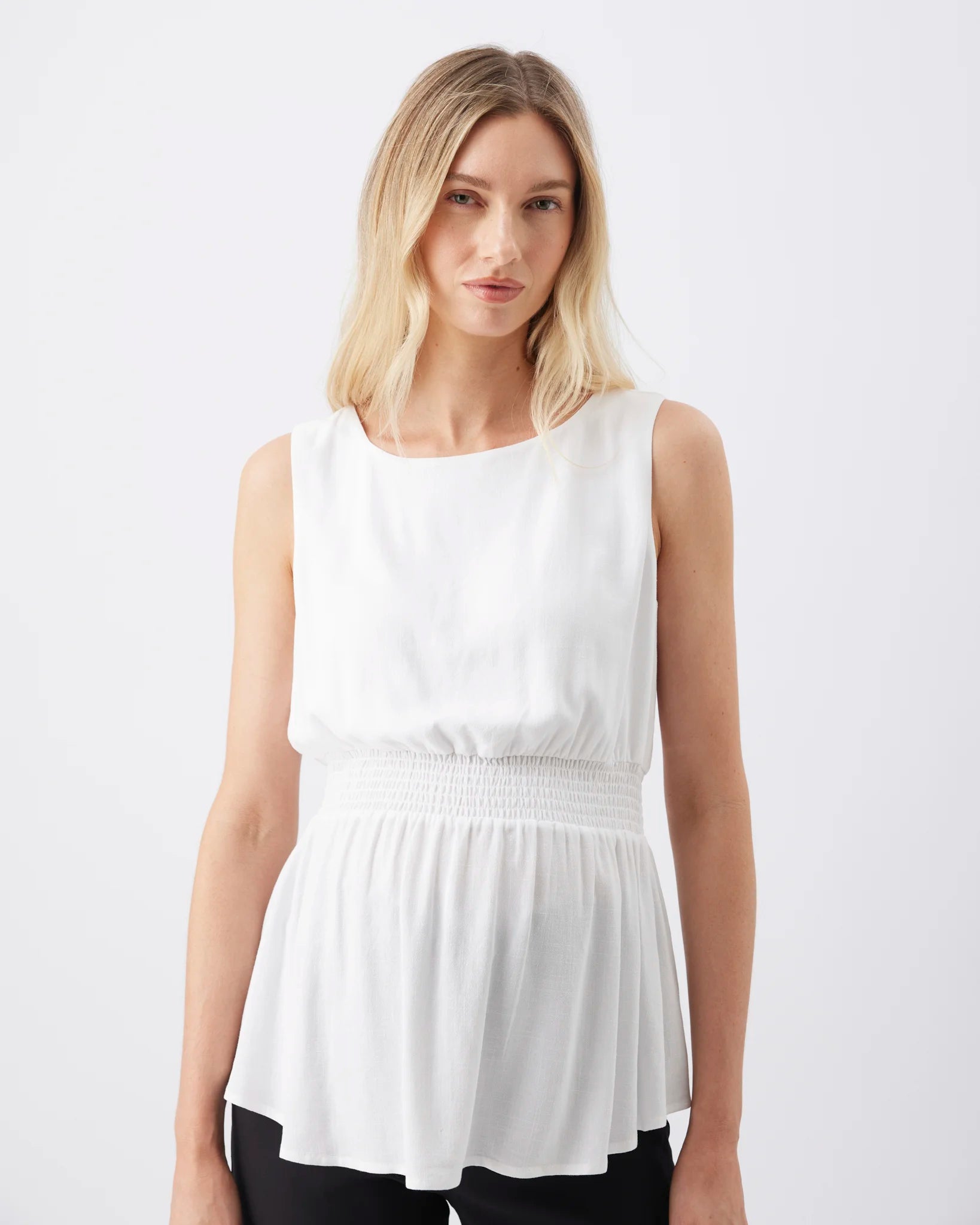 A blonde woman wearing a white sleeveless peplum top with a smocked waistband, paired with black pants, against a plain white studio background.