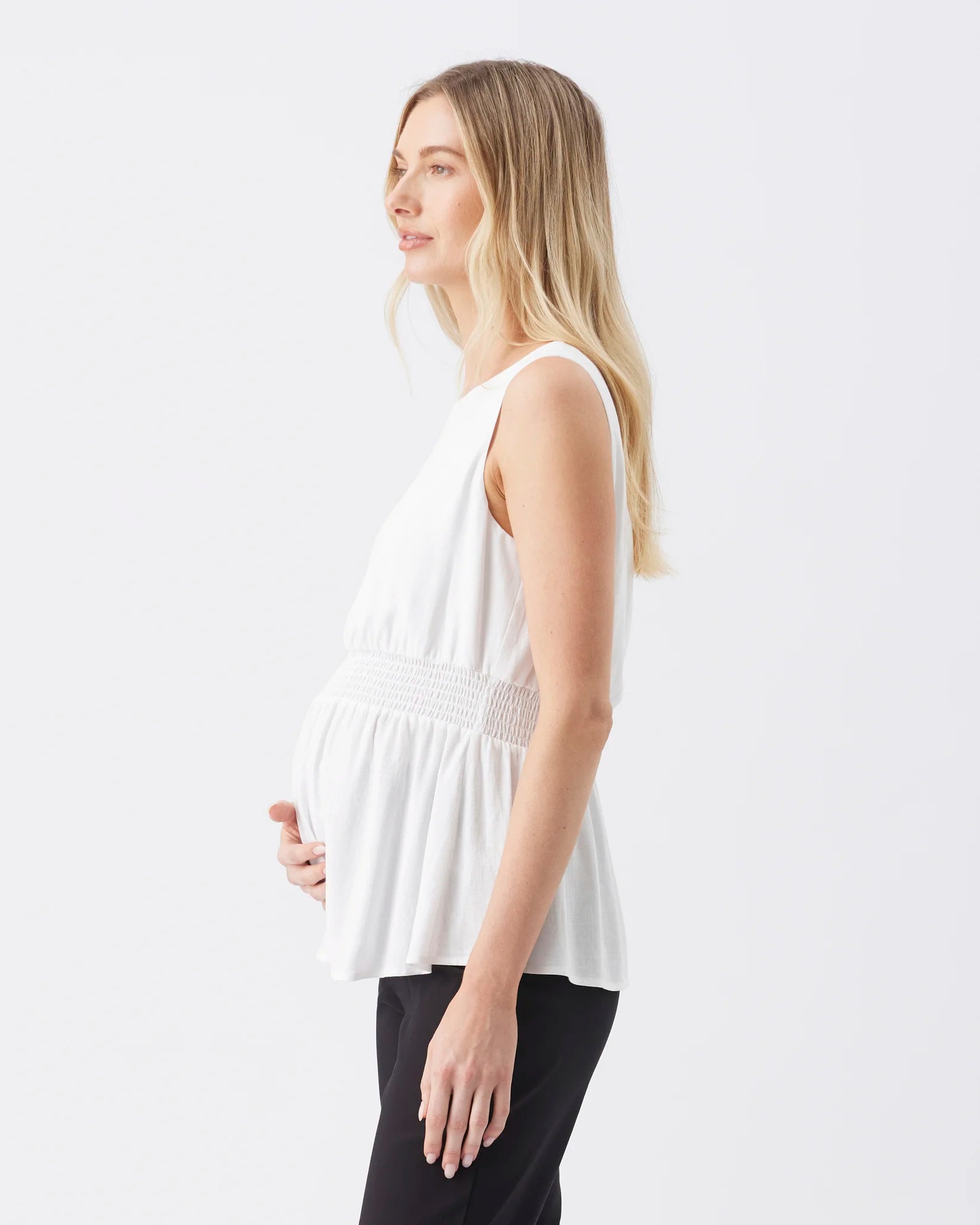 A pregnant woman in profile wearing a white sleeveless peplum top with a smocked waist and black pants, photographed against a clean white studio background.