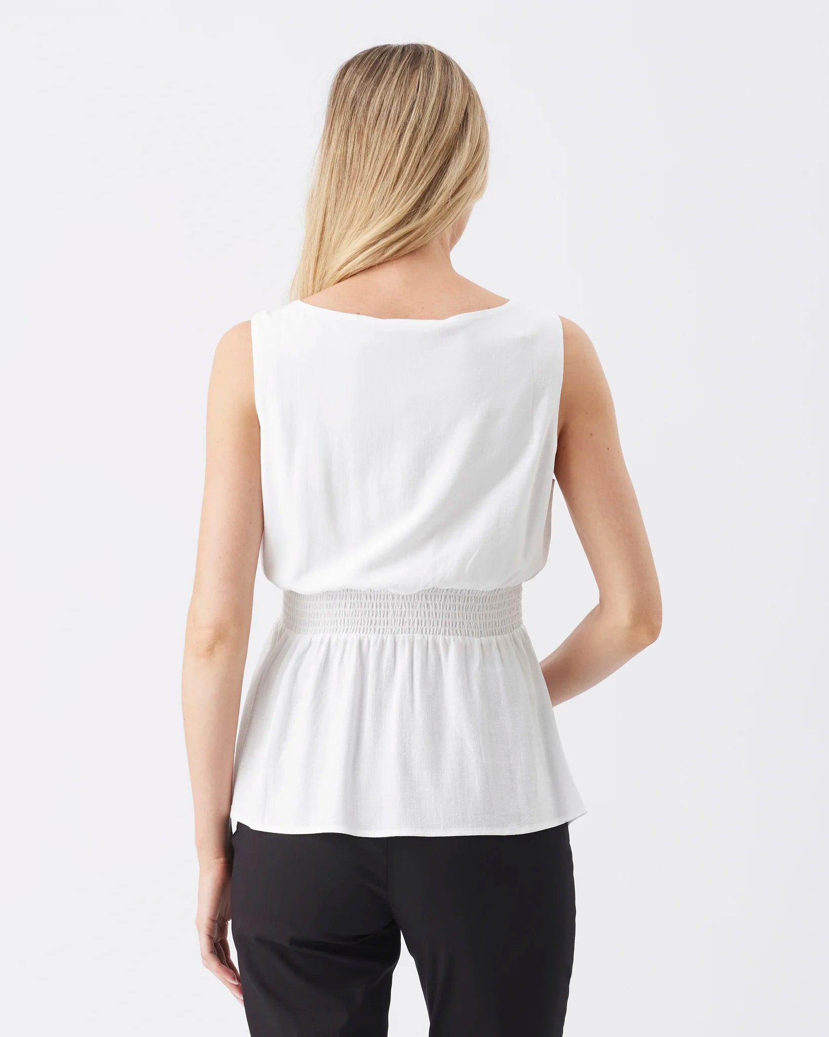 A woman shown from the back wearing a white sleeveless peplum top with a smocked waistband and black pants, against a light gray studio background.