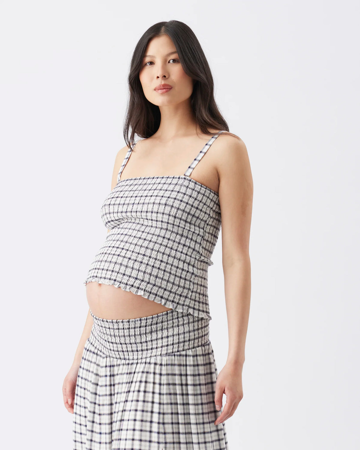 A pregnant model wearing a black and white gingham checkered maternity crop top and matching high-waisted maxi skirt, photographed against a clean white studio background.