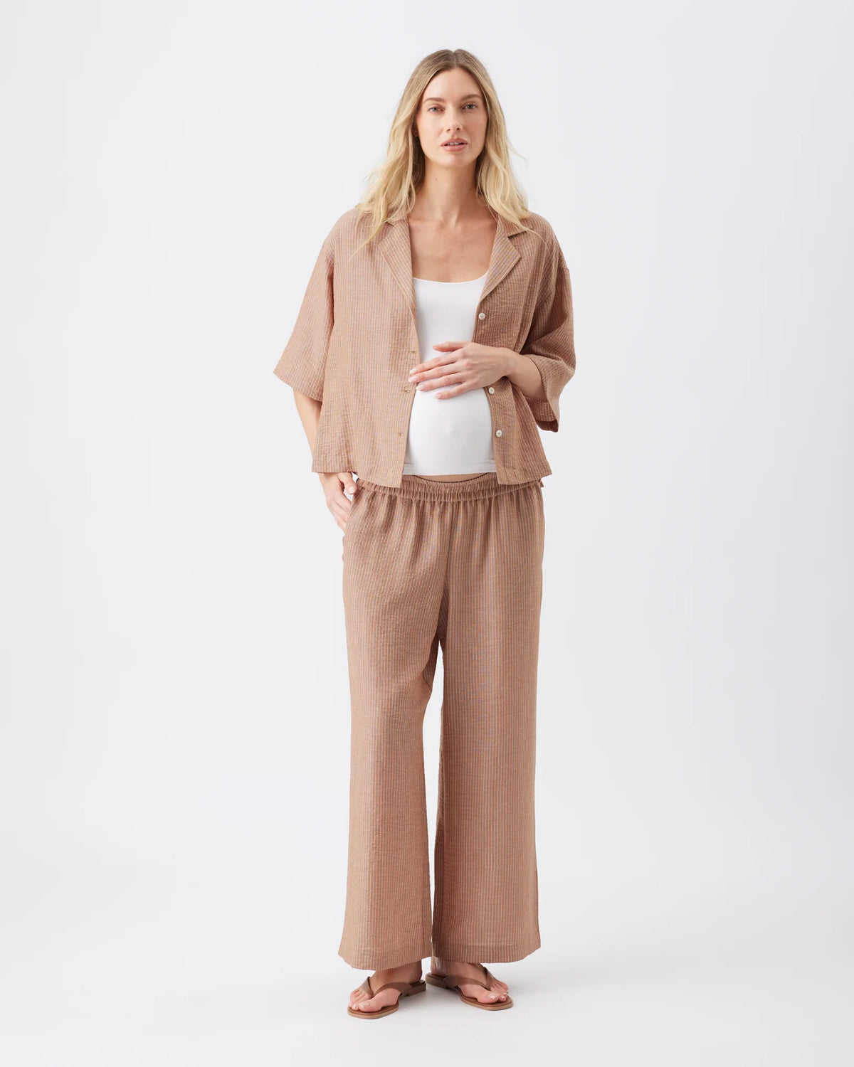 A pregnant woman modeling a matching beige linen two-piece set consisting of a short-sleeve button-up shirt and wide-leg trousers, styled over a white tank top and sandals against a plain white studio background.