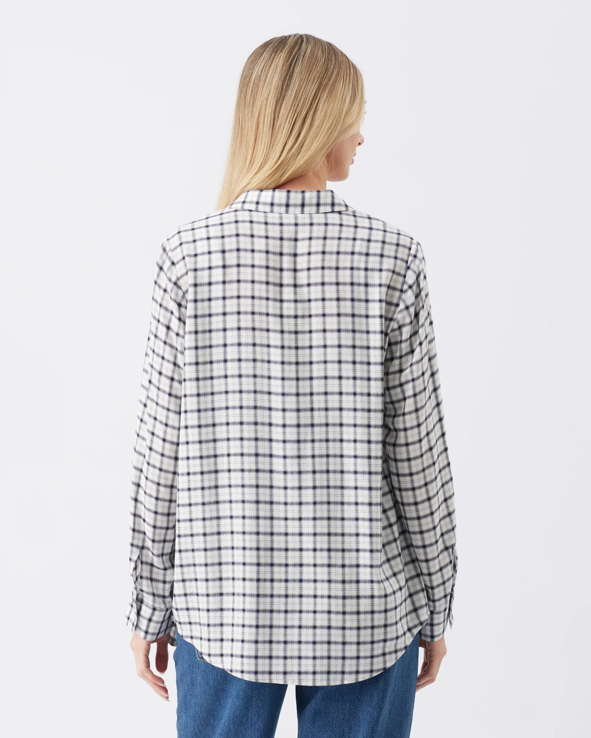A woman viewed from the back wearing a white long-sleeve button-up shirt with a black grid plaid pattern and blue jeans, set against a plain light gray studio background.