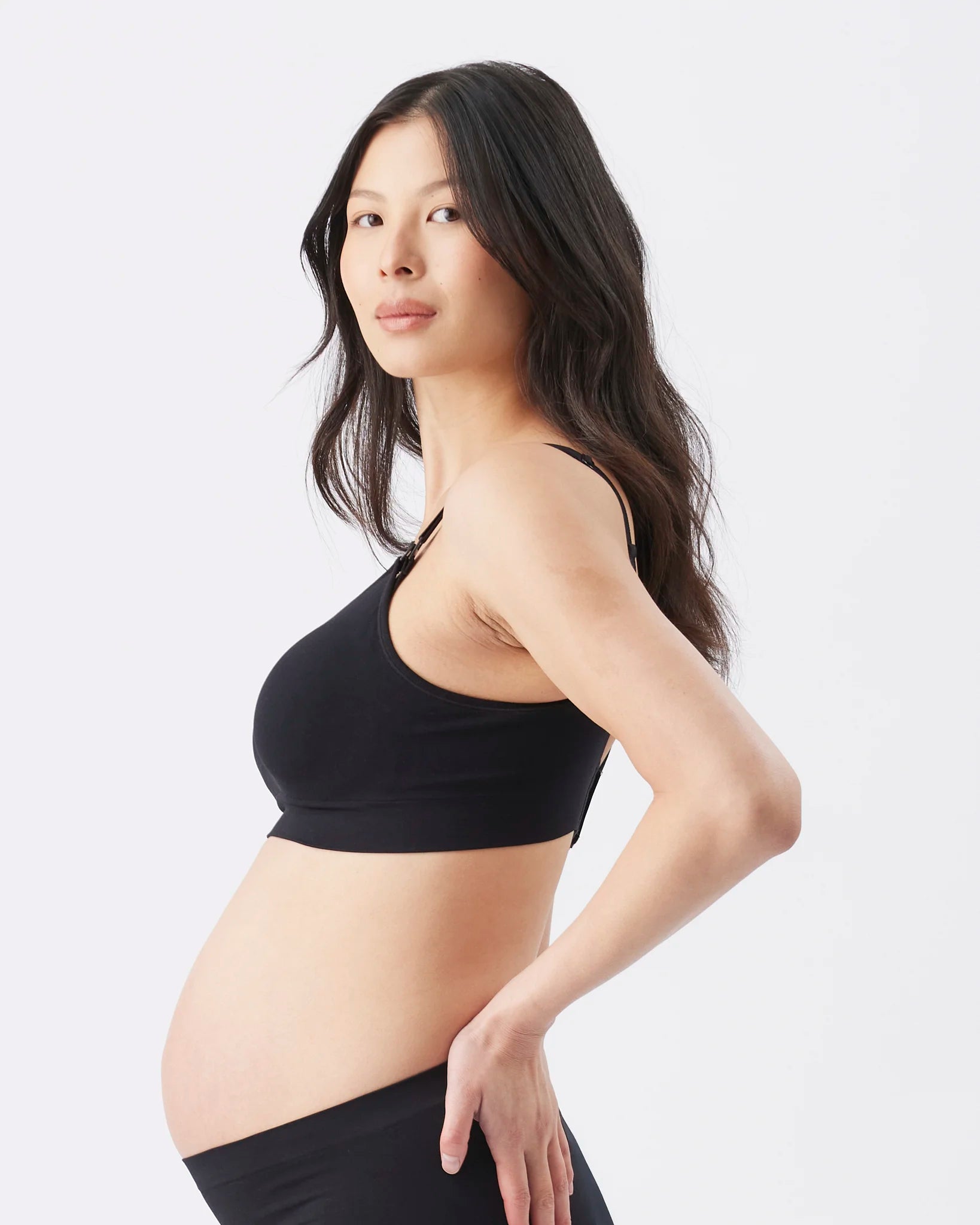 A pregnant woman in profile wearing a black maternity sports bra and black bottoms, photographed against a clean light gray studio background.
