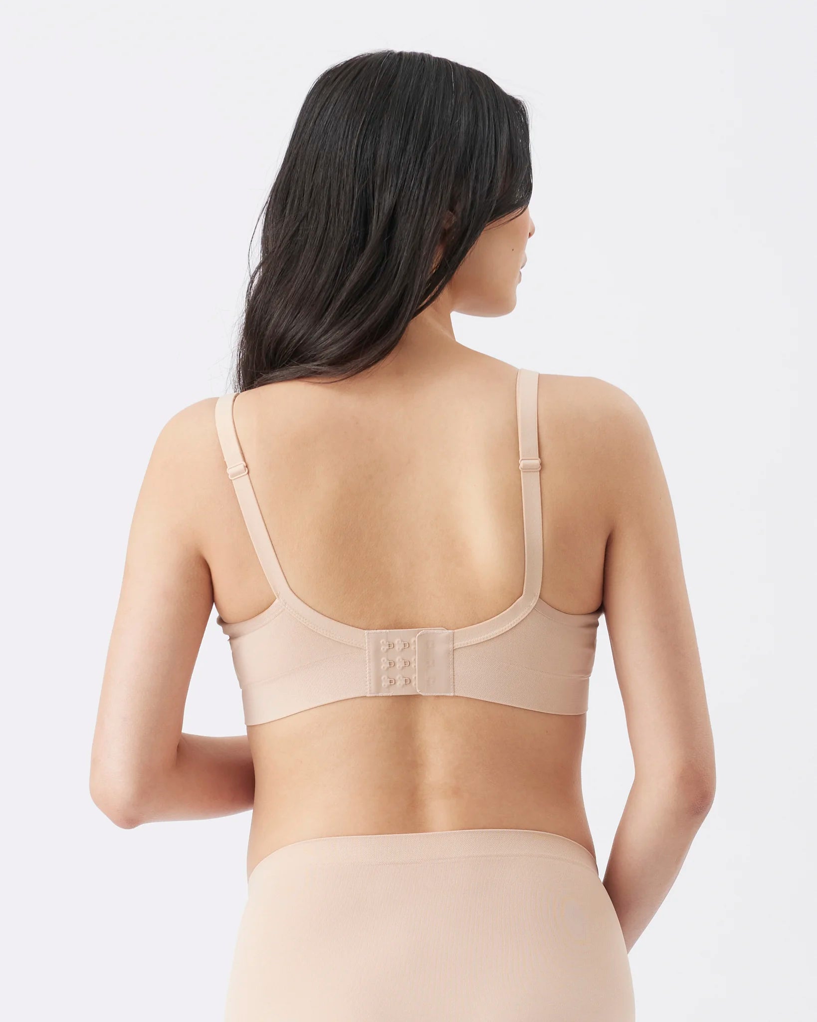 A woman shown from the back wearing a light beige bra and matching shapewear bottoms against a plain light gray studio background.