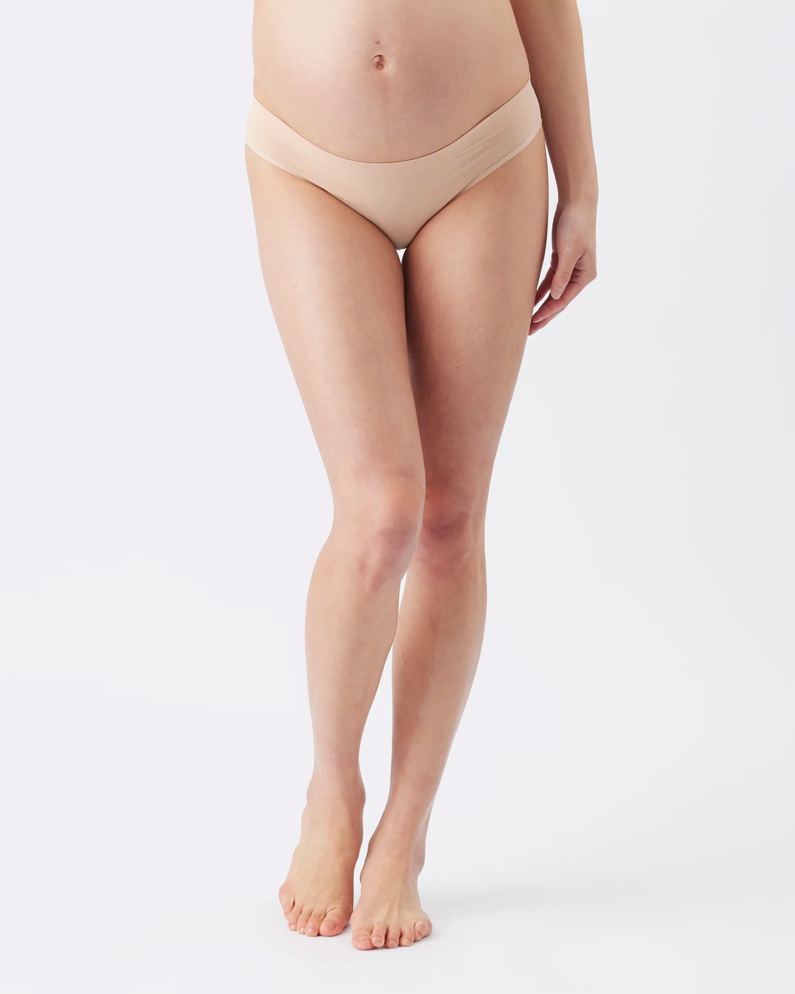 A model wearing a pair of high-waisted, low-rise maternity briefs in a nude beige color, shown from the waist down against a clean white studio background.