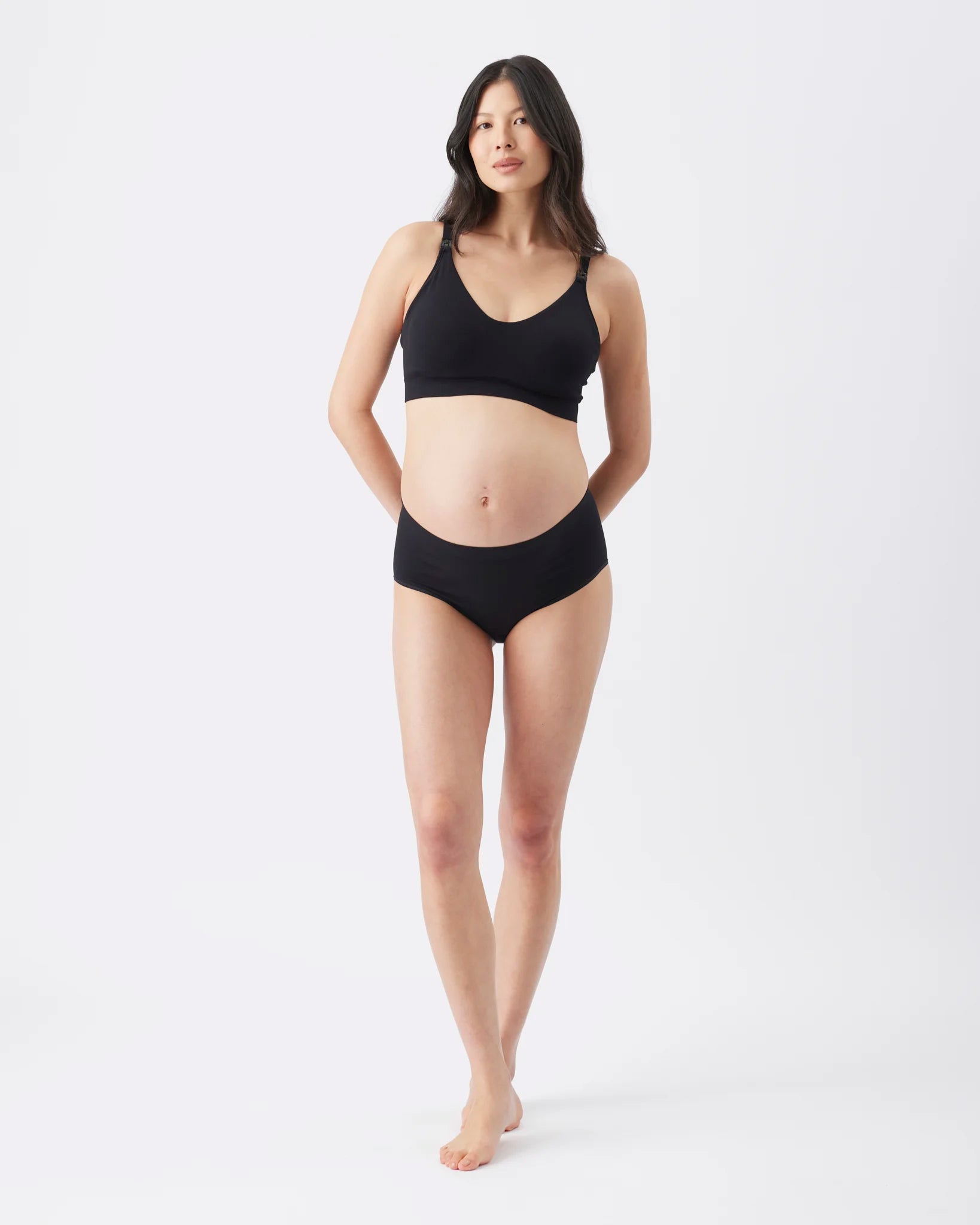 A pregnant model wearing a black maternity bra and matching black underwear, standing against a seamless white studio background.