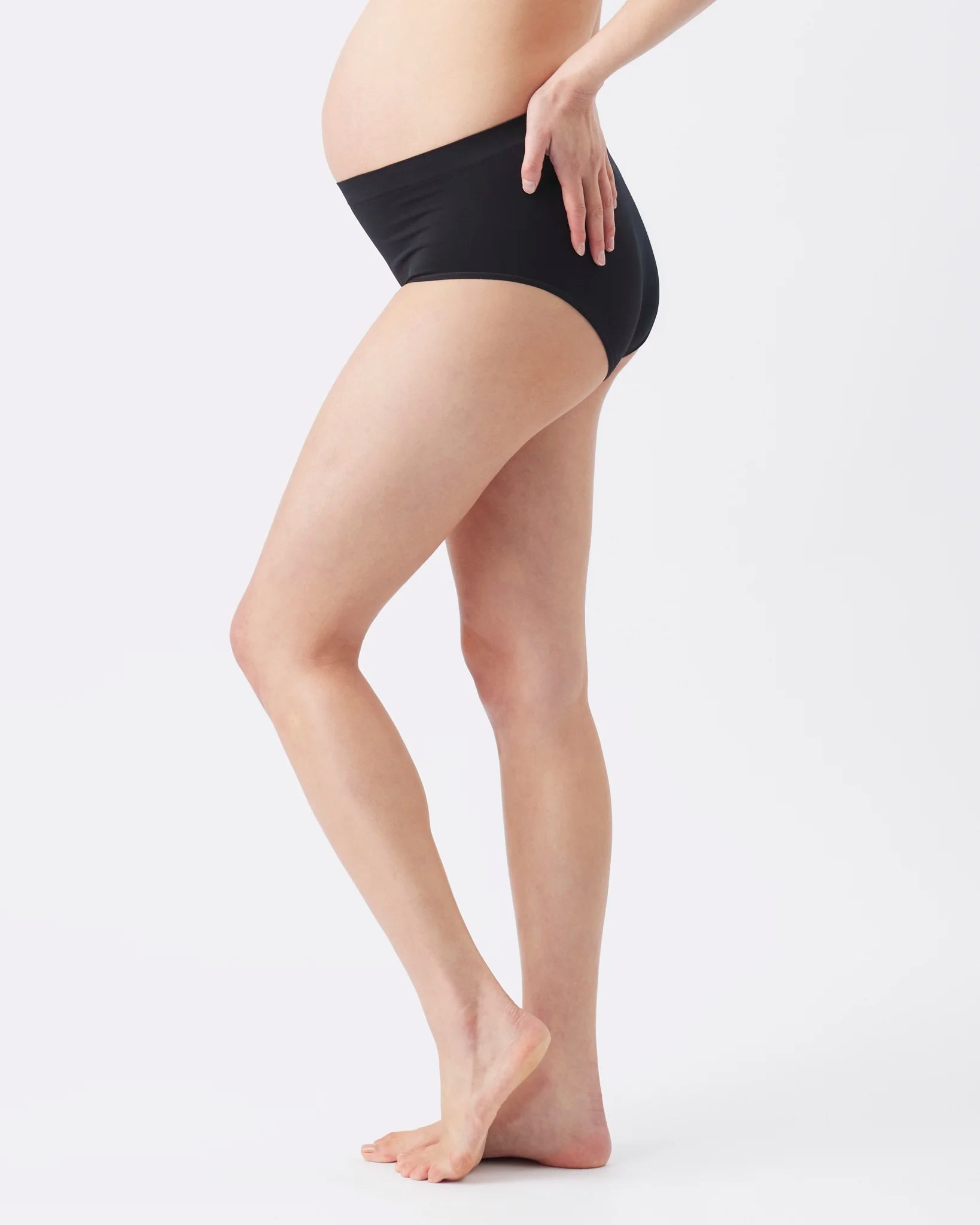 A side-profile view of a pregnant model wearing black maternity underwear, standing barefoot against a clean white studio background.