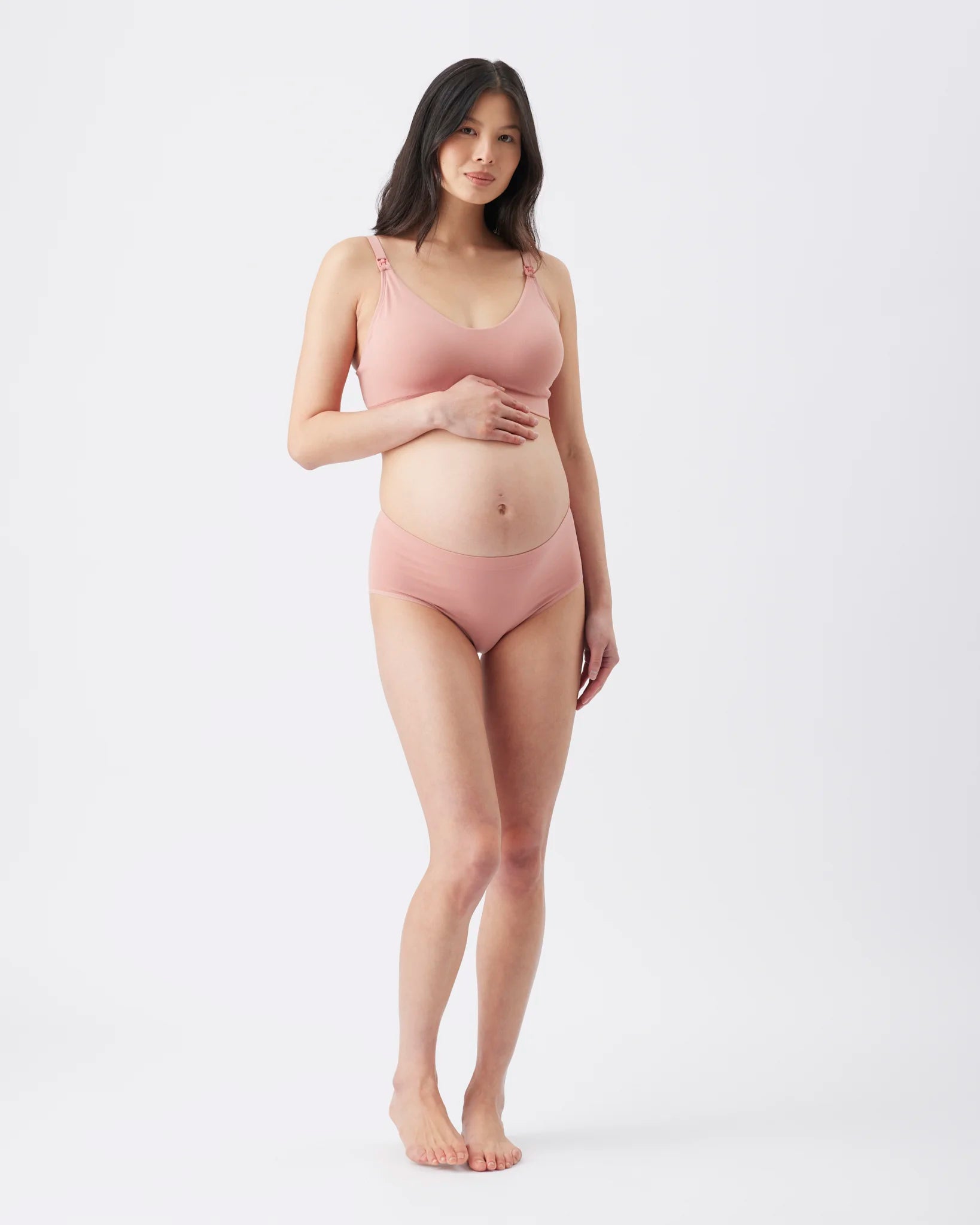 A pregnant woman modeling a matching dusty pink maternity bra and underwear set against a plain white studio background.