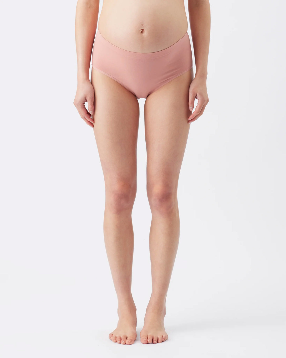 A model from the waist down wearing a pair of high-waisted, seamless maternity briefs in a dusty rose pink color, standing against a plain white studio background.