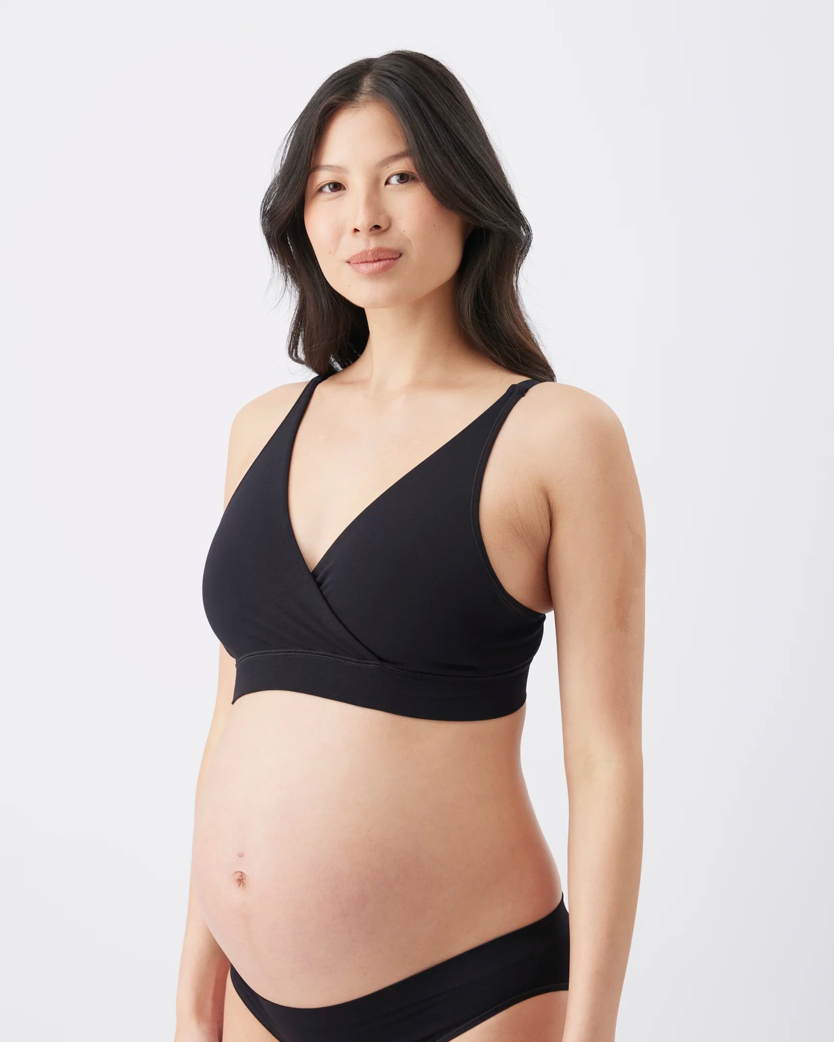 A pregnant woman wearing a black nursing bra and matching black underwear, standing against a plain light gray studio background.