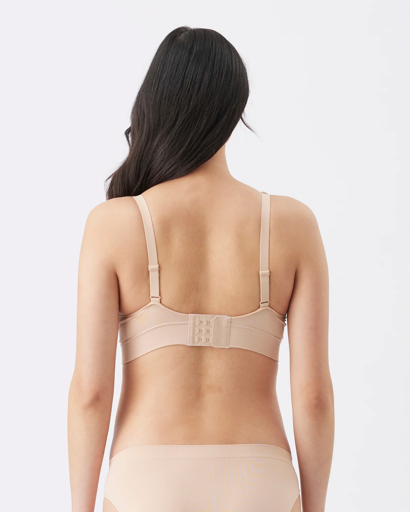 Rear view of a woman wearing a beige wireless bra and matching underwear against a light gray studio background.