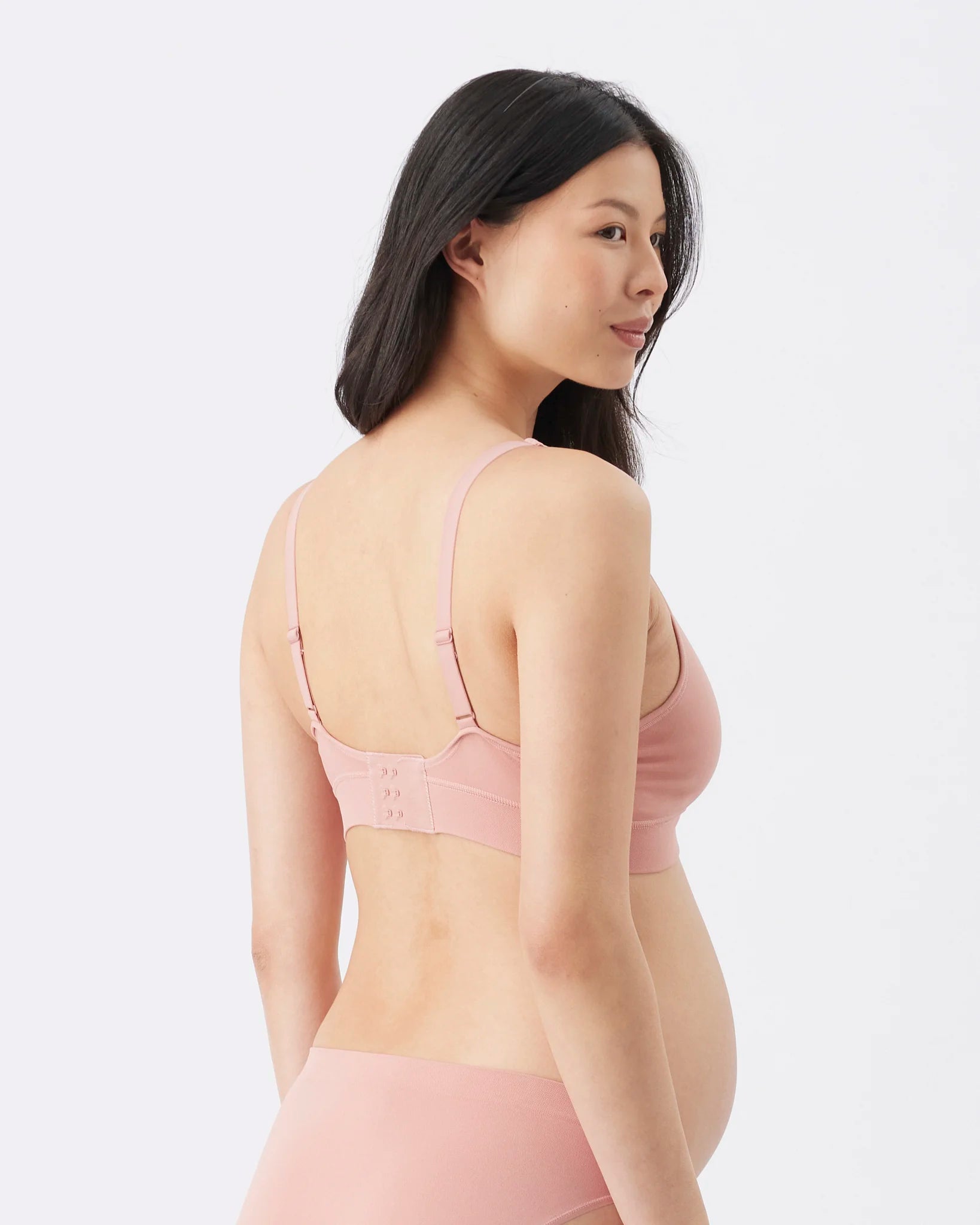 A pregnant model shown from the back wearing a light pink maternity bra and matching underwear against a plain white studio background.