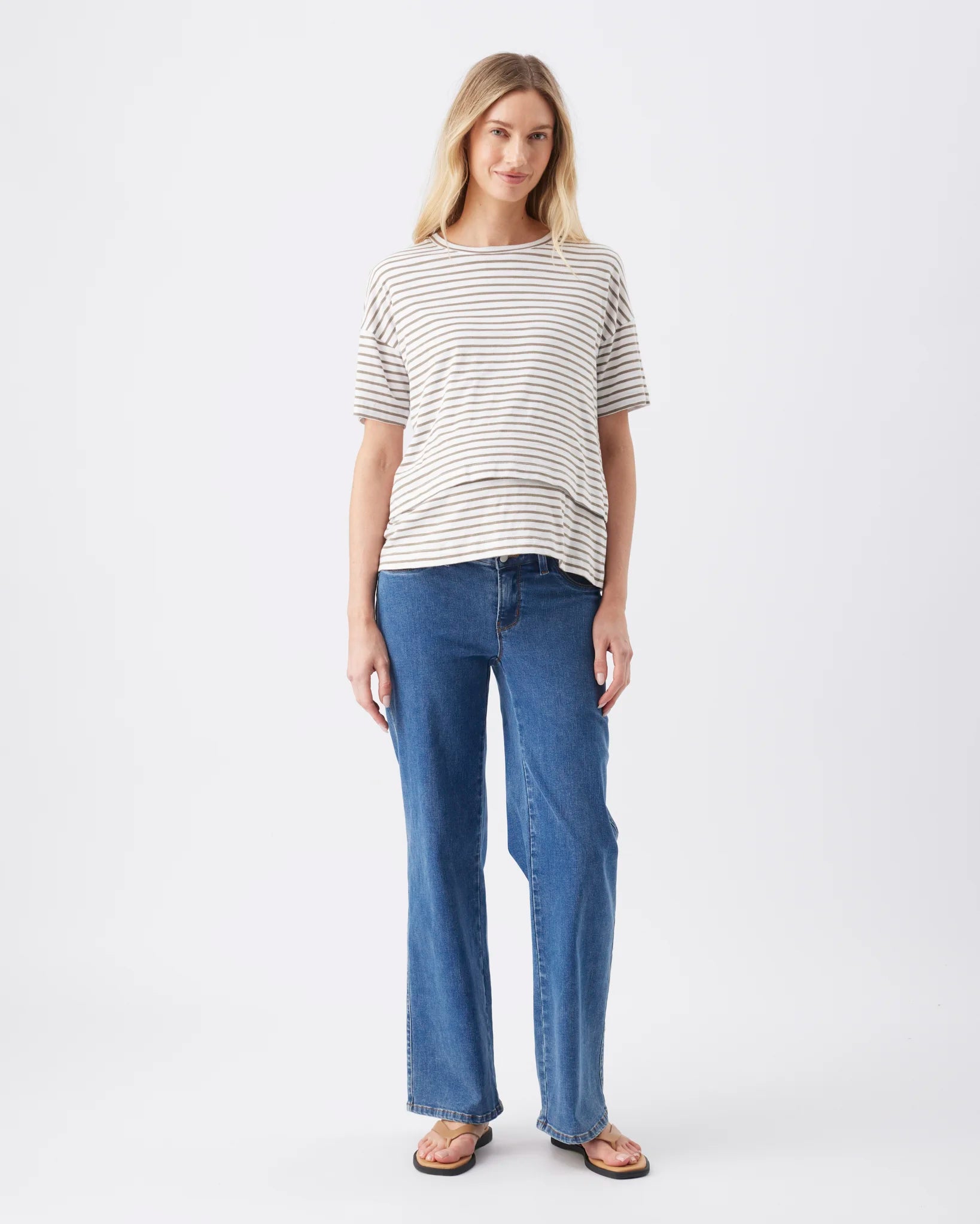 A woman modeling a short-sleeve crewneck t-shirt with thin dark horizontal stripes on a light beige background, paired with blue jeans and brown sandals, against a plain white studio background.
