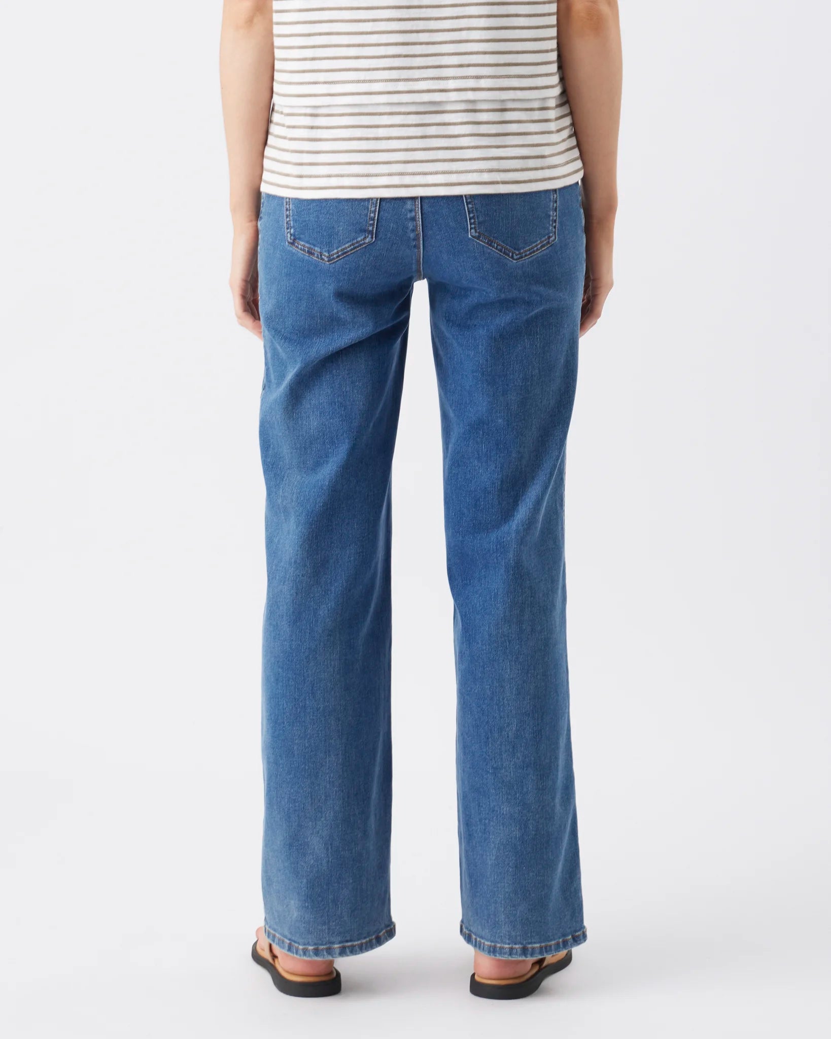 Rear view of a person wearing blue wide-leg jeans, a white striped tank top, and brown sandals against a light gray studio background.