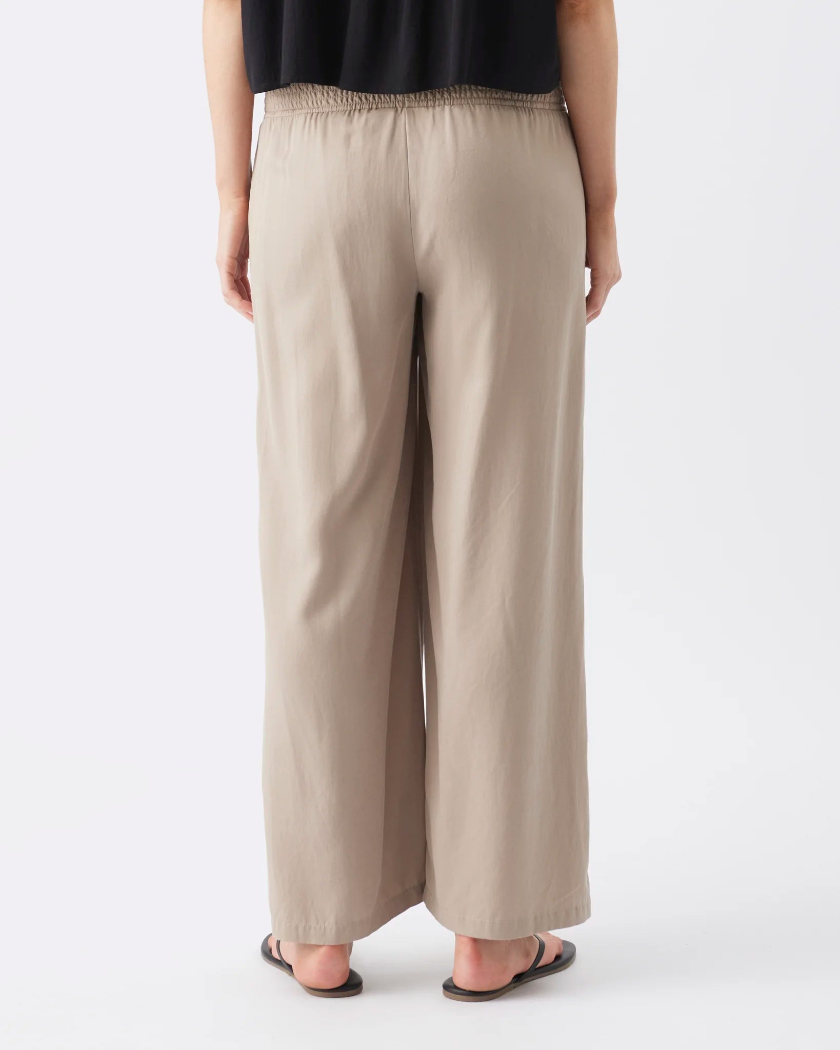 A person shown from the back wearing loose, wide-leg beige pants with an elastic waistband, paired with a black top and dark sandals, against a plain light gray studio background.