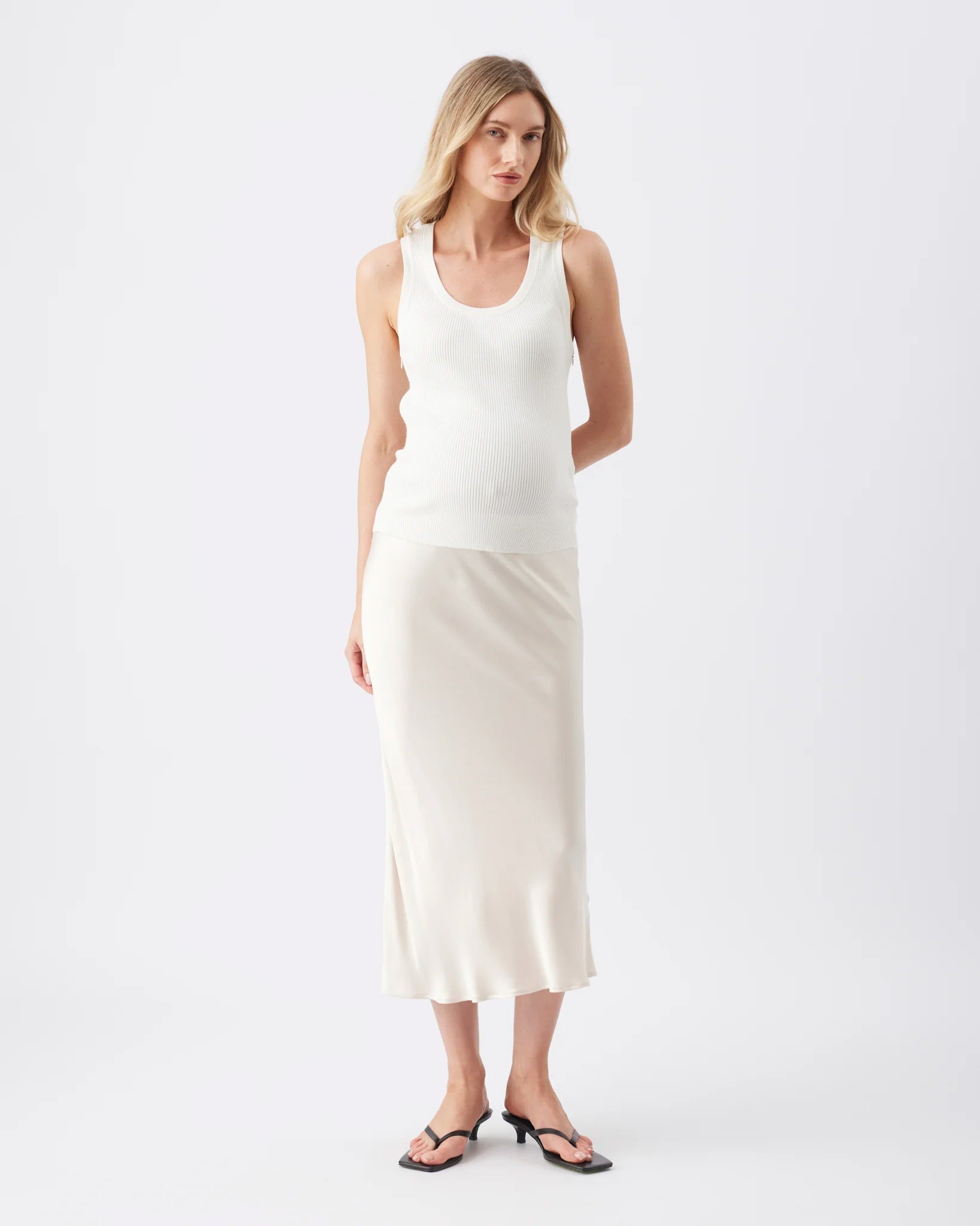 A woman modeling a sleeveless white ribbed knit tank top and a matching white midi skirt, paired with black thong sandals, against a plain white studio background.