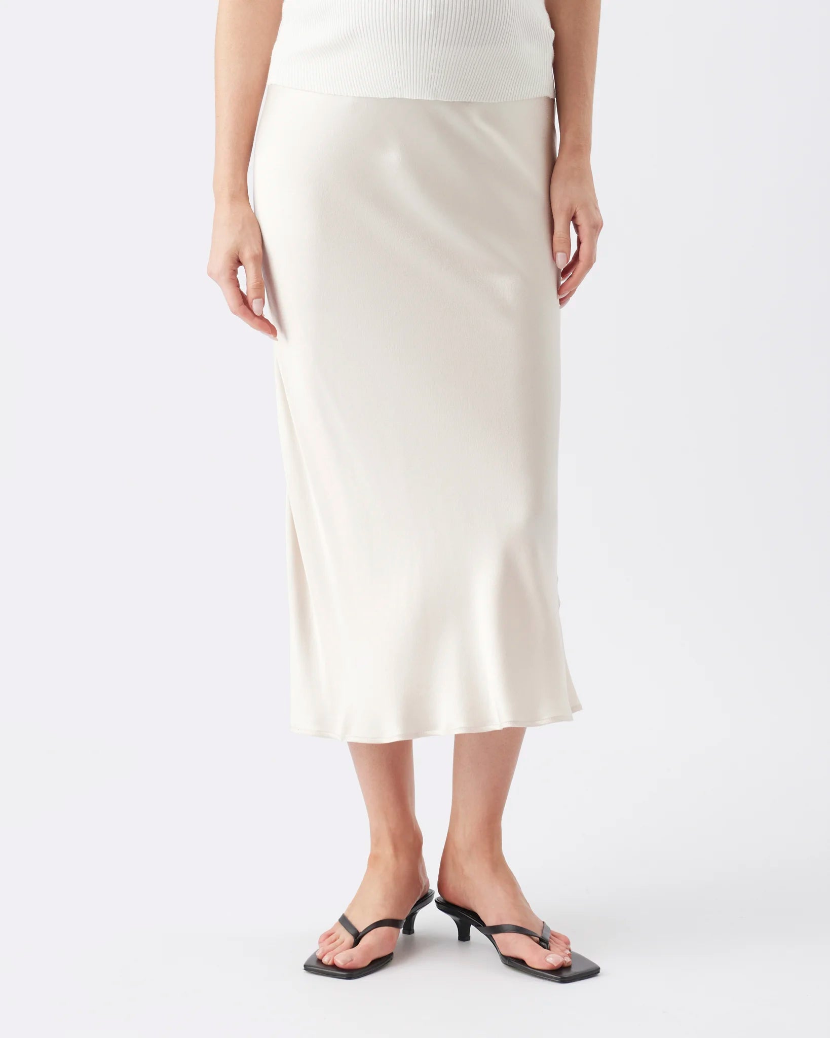 A model wearing a cream-colored satin midi skirt with a side slit, paired with black thong sandals and a white ribbed top, against a plain light gray background.
