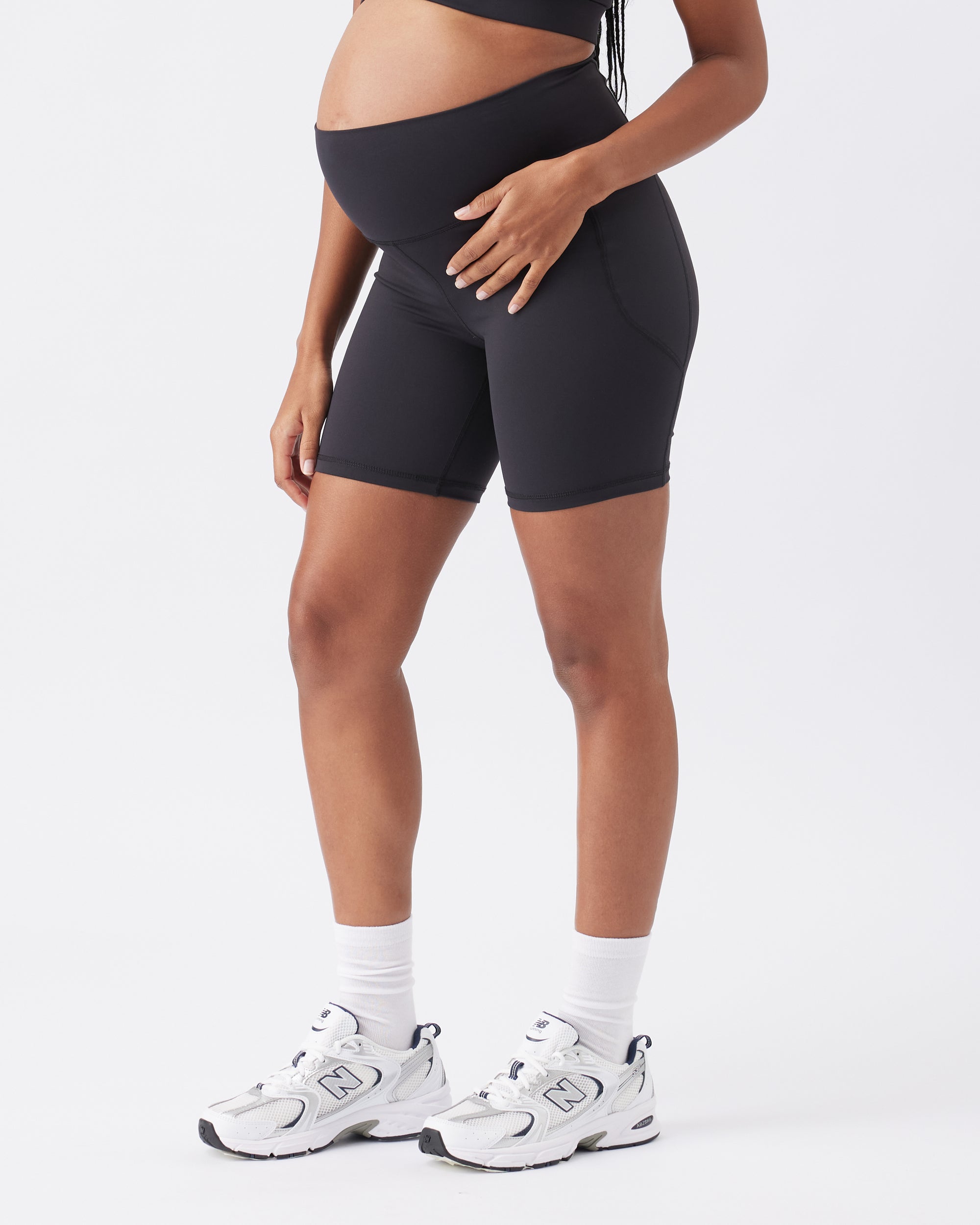 Pregnant woman wearing black bike shorts and white sneakers on a white background