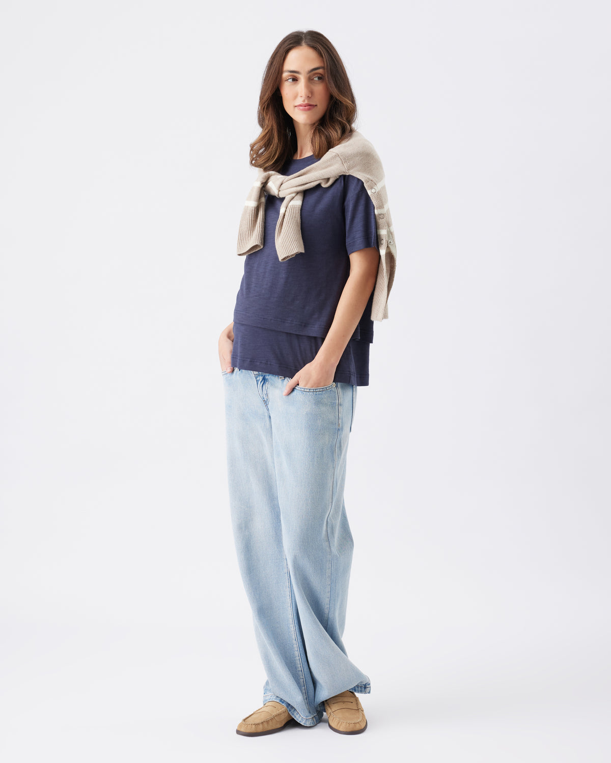 Woman wearing a navy top, light blue jeans, and a beige jumper tie dover the shoulders on a white background