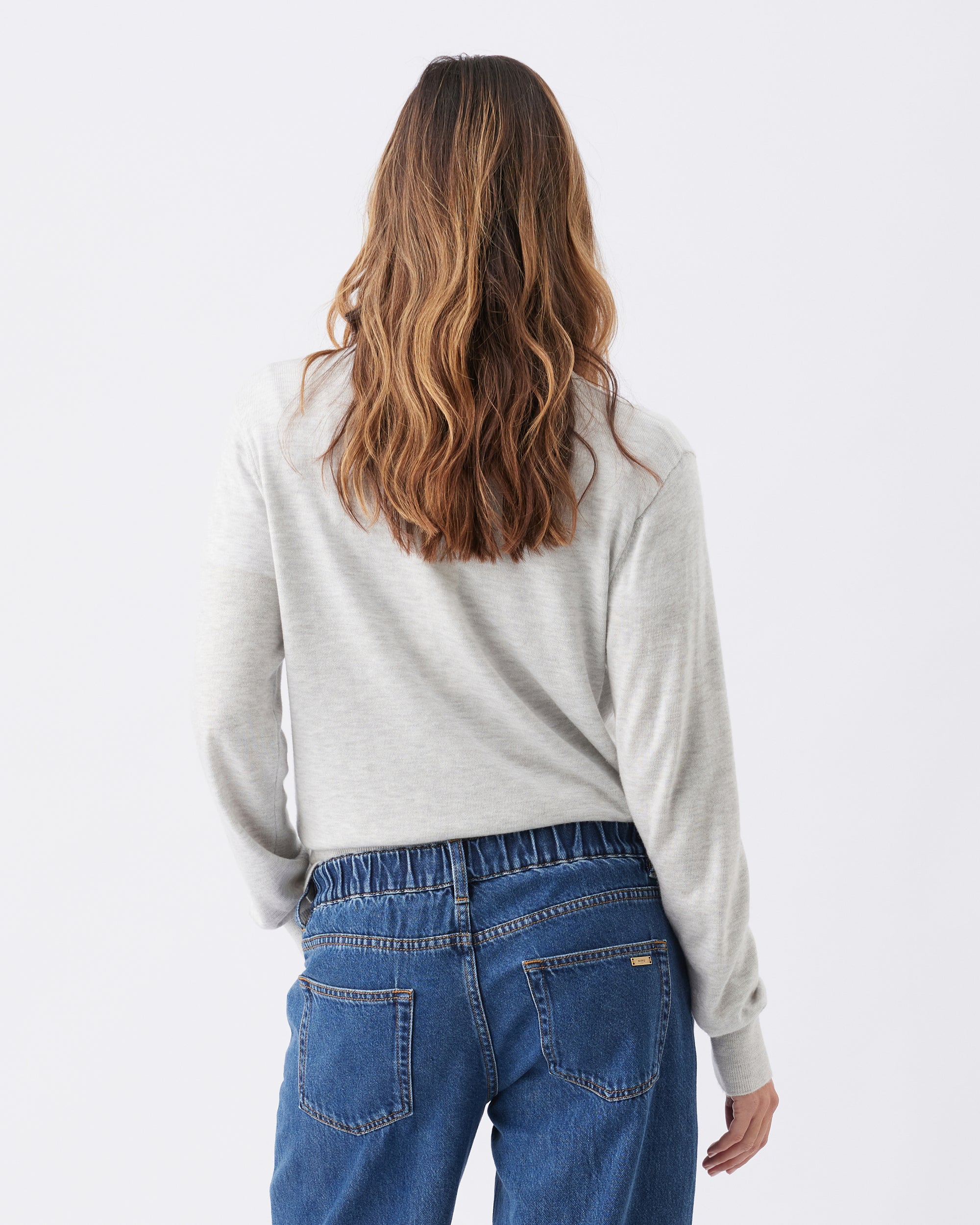 Back view of a woman wearing a gray long sleeve top and blue jeans on a white background