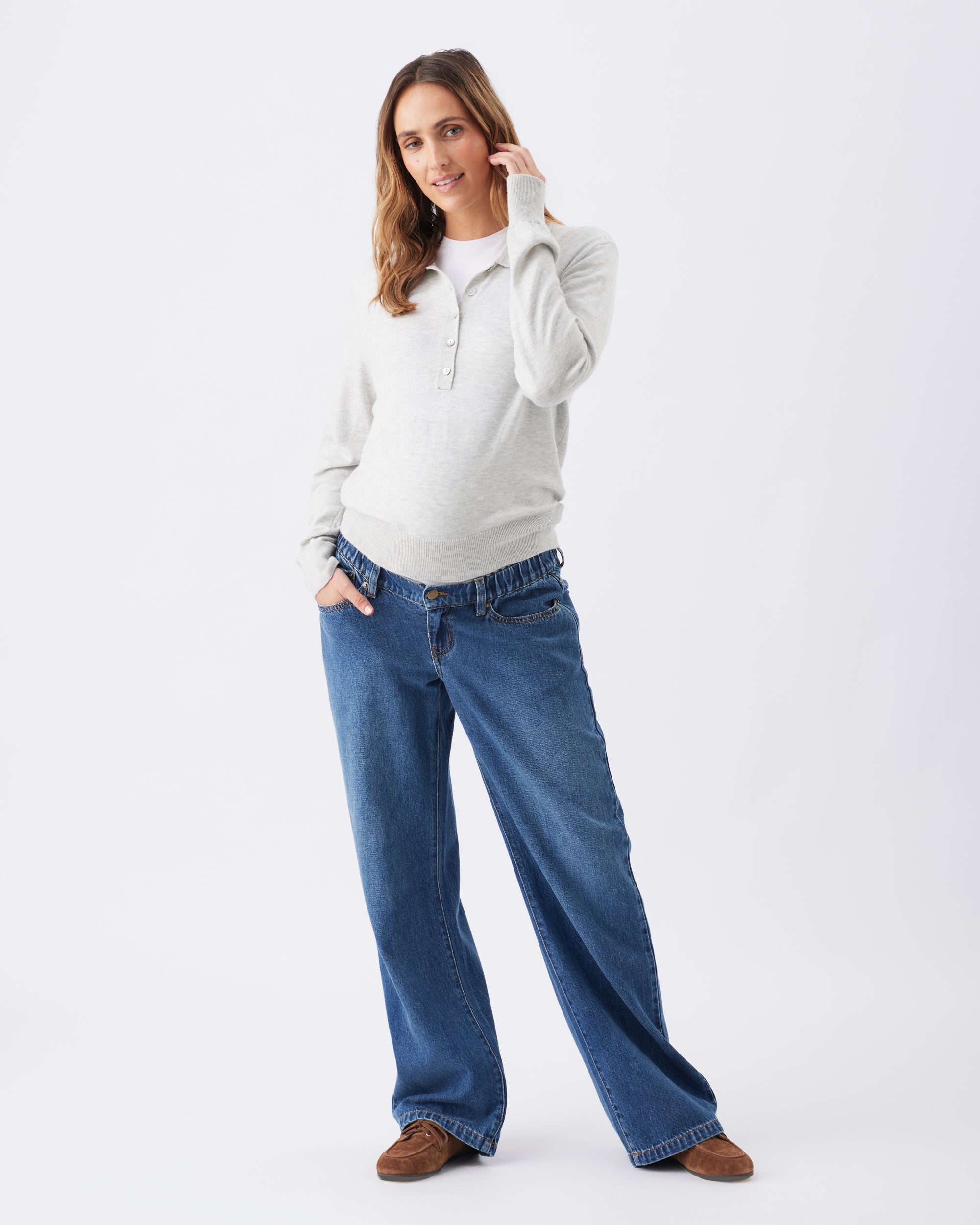 Pregnant woman wearing a light grey knitted top and dark blue maternity jeans on a white background