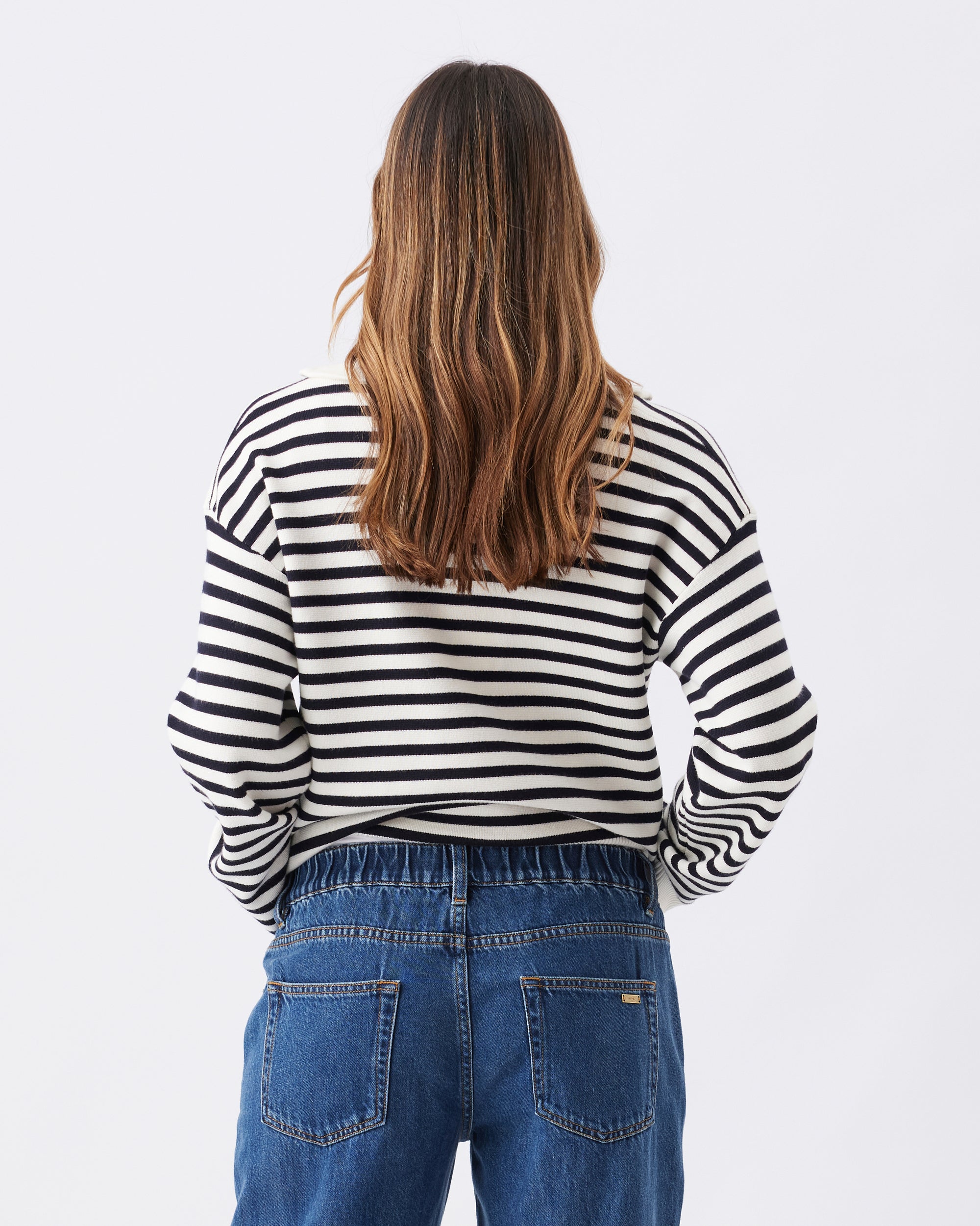 Back view of a woman wearing a dark blue and white striped top and blue jeans on a plain background