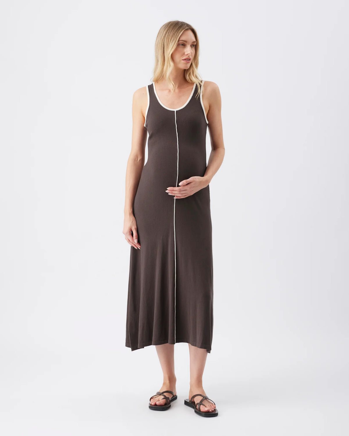 A pregnant model wearing a dark brown maternity tank dress with white trim and a front zipper, paired with black sandals, against a plain white studio background.