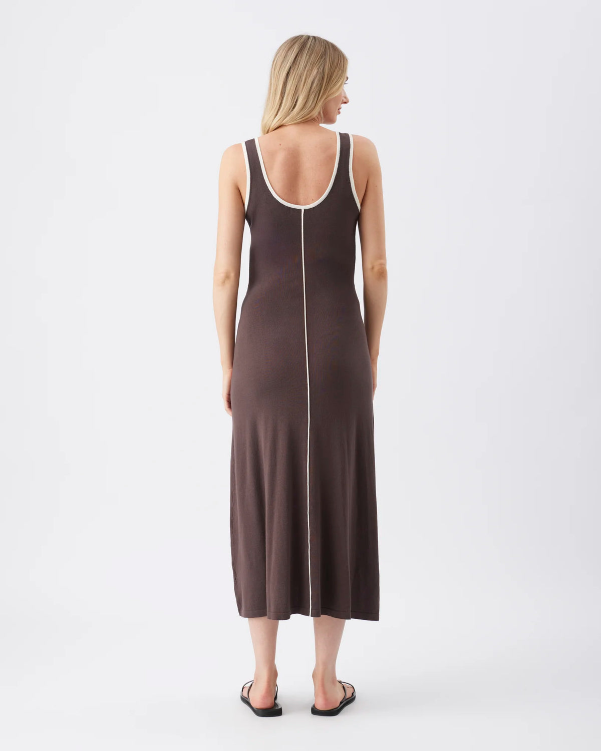 A woman viewed from the back wearing a sleeveless midi dress in a dark taupe-brown color with a contrasting white trim along the scoop neckline and a vertical white seam down the center back. She is standing against a plain white studio background.