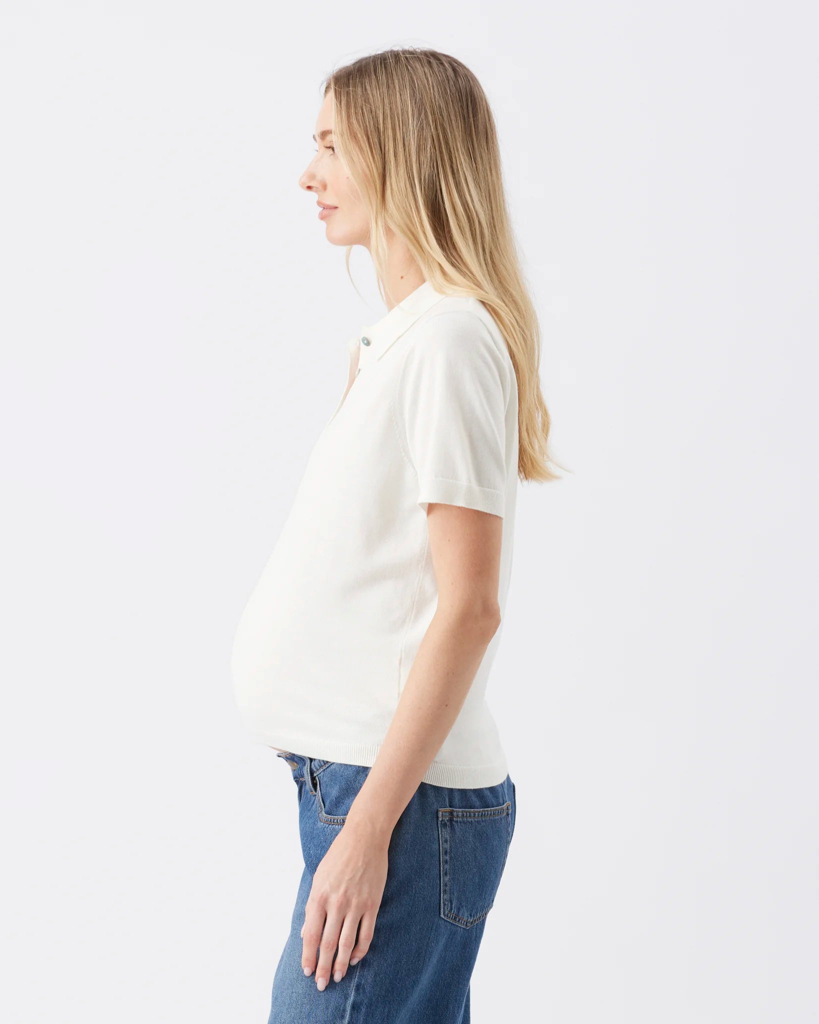 A pregnant woman shown in profile wearing a white short-sleeve polo shirt and blue jeans against a plain white studio background.