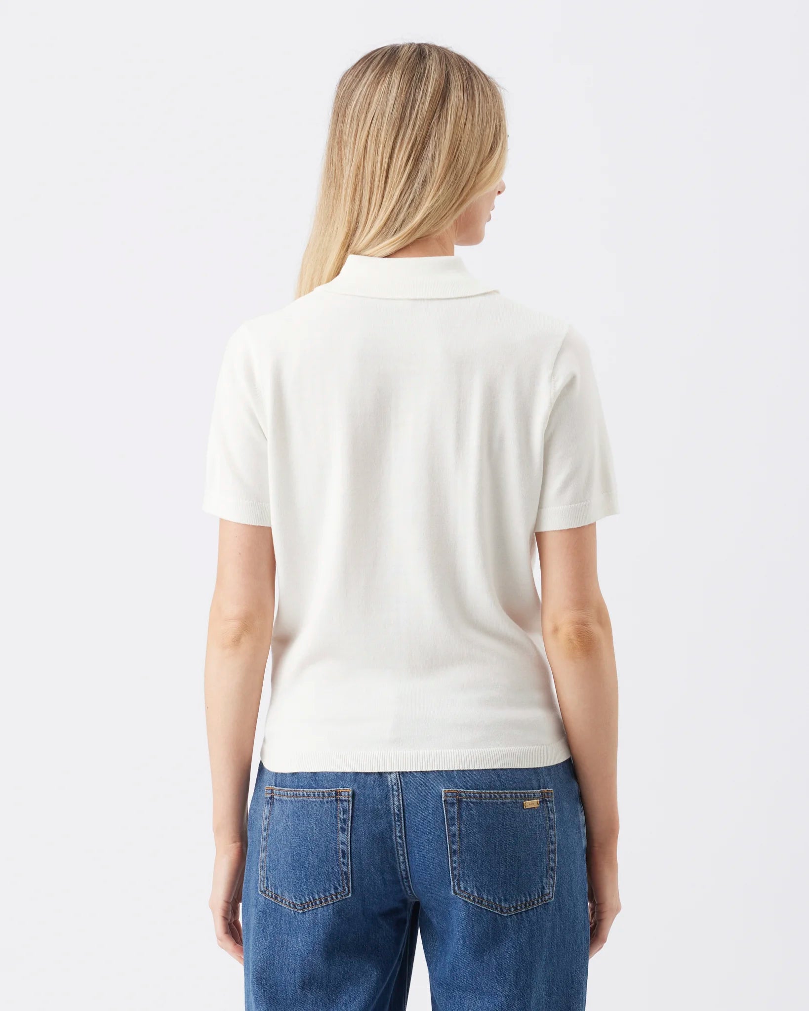 A woman viewed from the back wearing a short-sleeve white polo shirt and blue jeans against a plain light gray studio background.