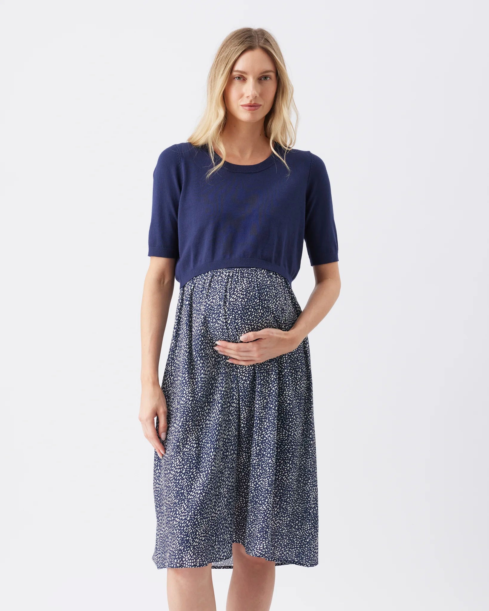 A pregnant woman modeling a maternity dress with a solid navy blue short-sleeve top and a navy and white speckled patterned skirt, set against a plain light gray background.