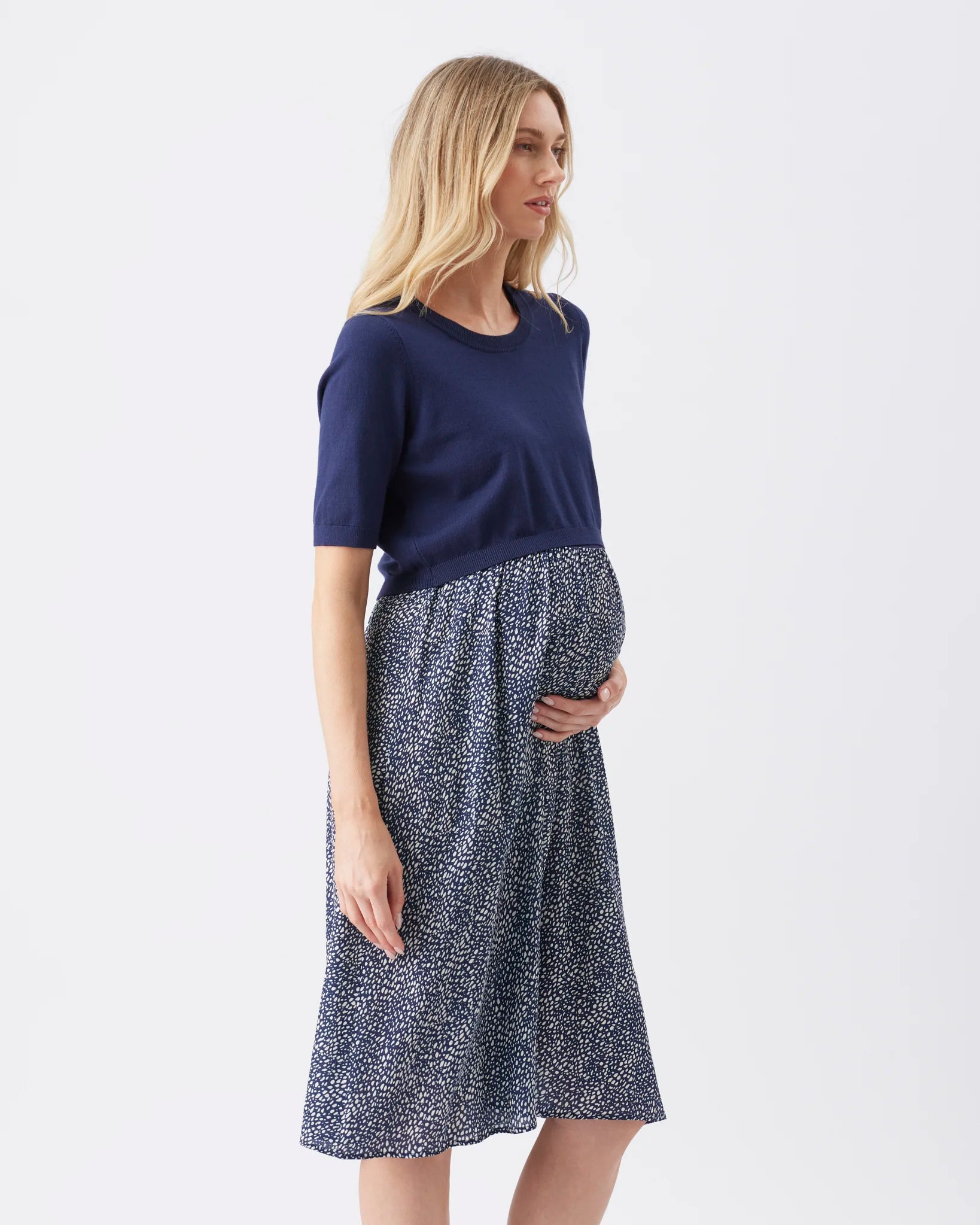 A pregnant woman wearing a navy blue short-sleeve top and a matching navy and white patterned maternity skirt, standing against a plain white studio background.