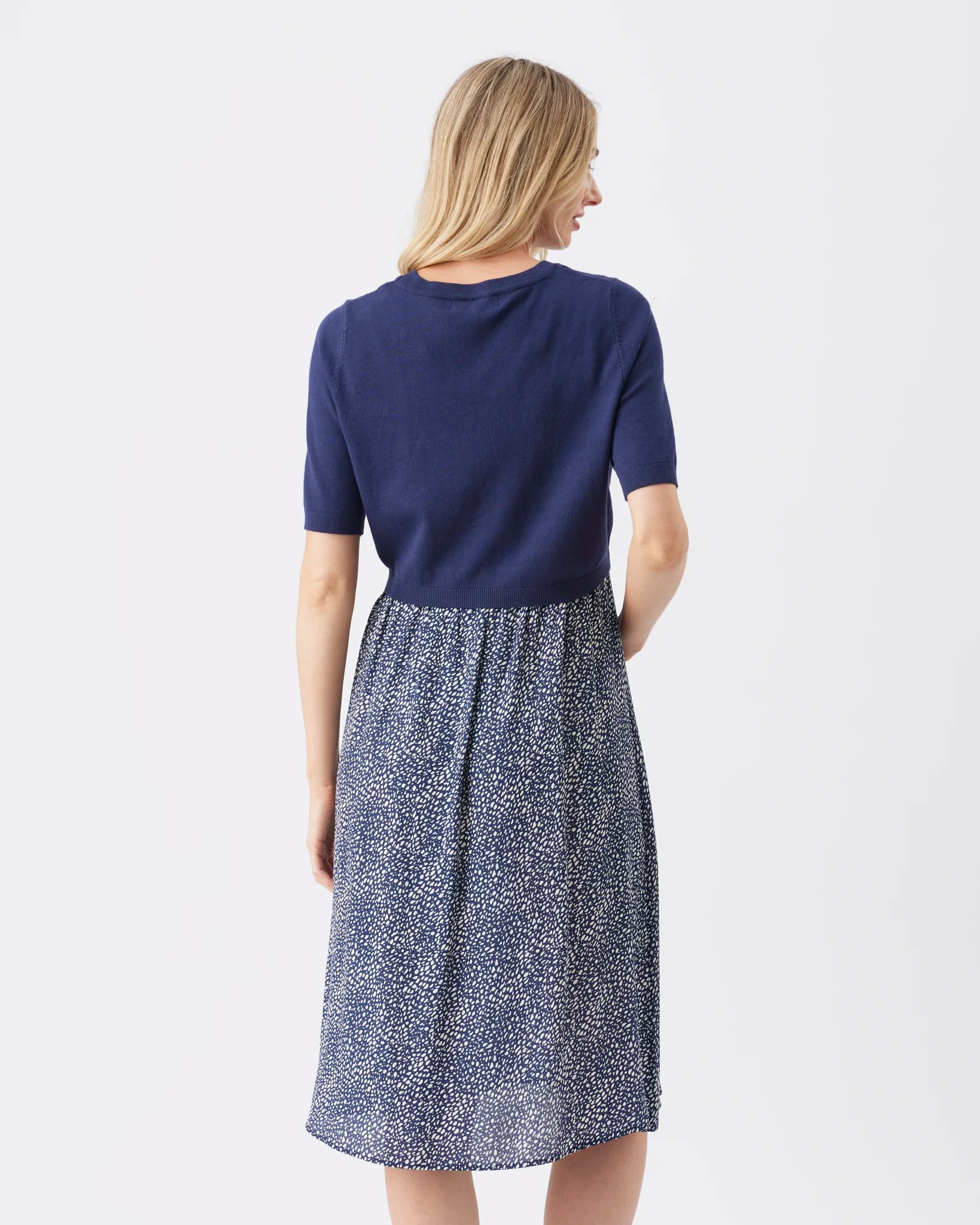 A woman shown from the back wearing a short-sleeve knit dress with a solid navy blue upper section and a navy and white speckled patterned skirt, against a plain white studio background.