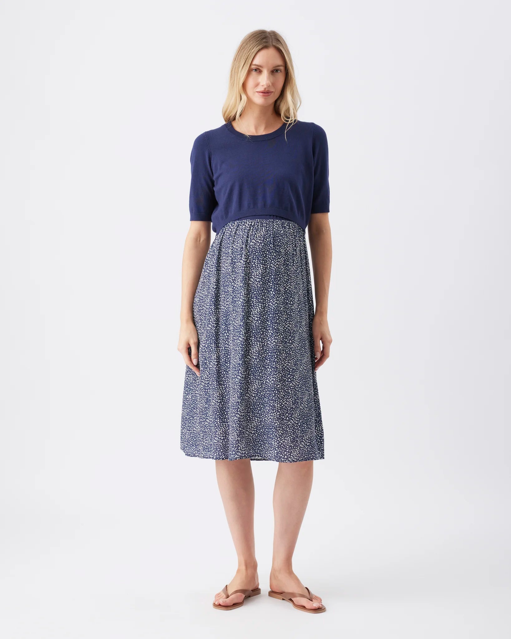 A woman modeling a short-sleeve knit dress with a solid navy blue top and a blue-and-white abstract patterned skirt, paired with tan flip-flop sandals against a light gray studio background.